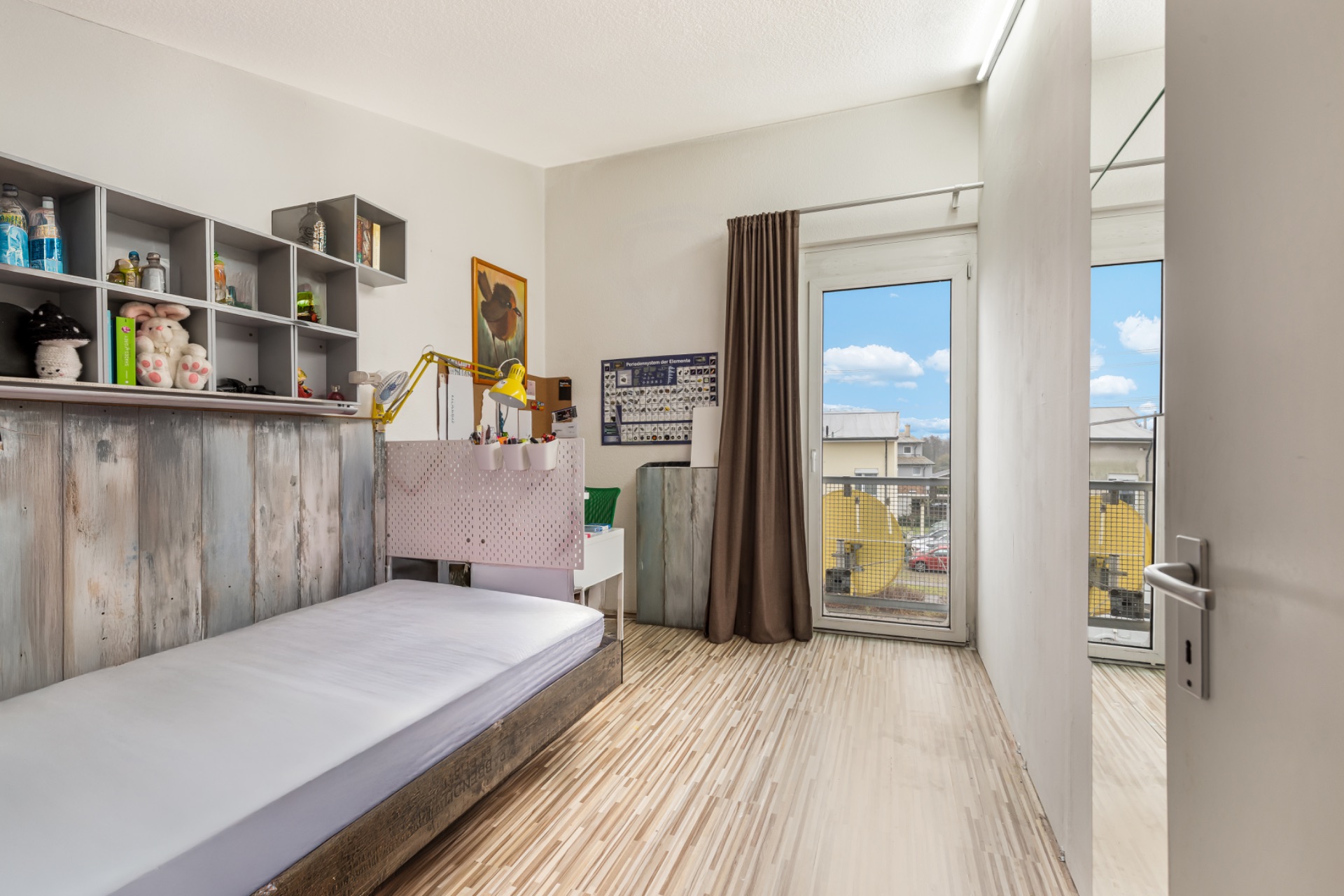 Helles Kinderzimmer mit Einzelbett, Regal, Schreibtisch und Fenster mit Vorhang.
