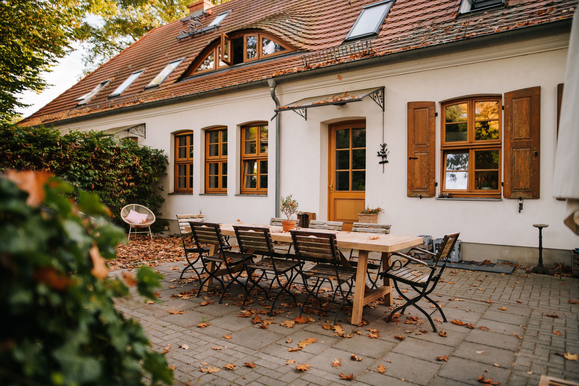 Großer Holztisch mit Stühlen auf einer Terrasse vor einem weißen Haus mit Holzfenstern.