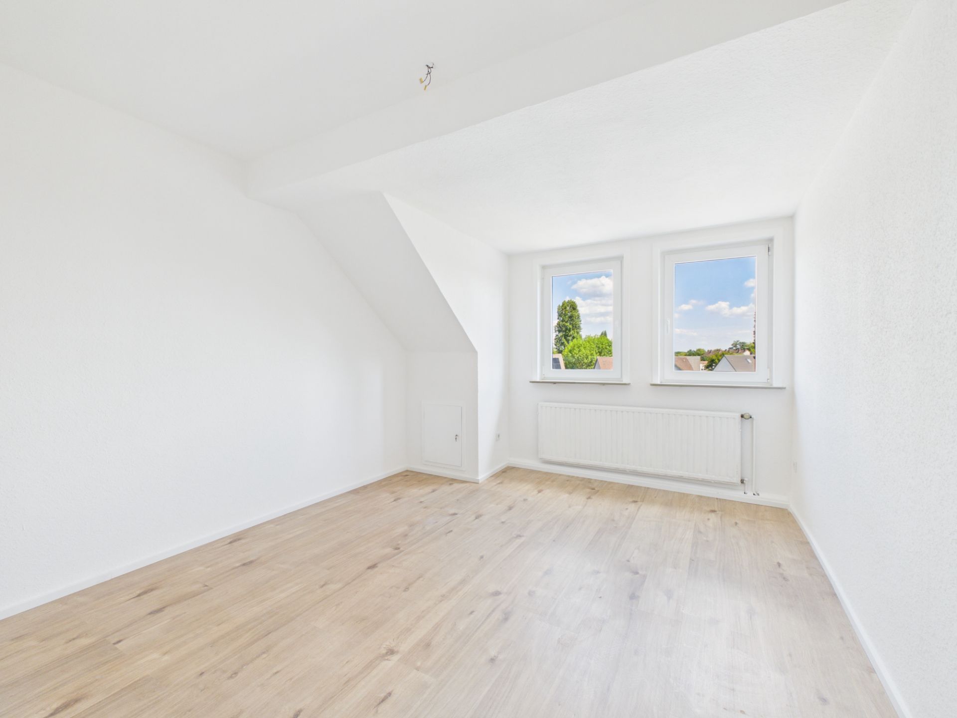 Helles, leeres Zimmer mit zwei Fenstern, weiß gestrichenen Wänden und hellem Holzfußboden.