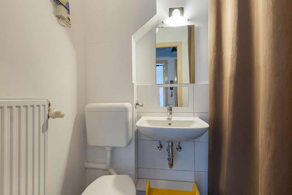Kleines Badezimmer mit Waschbecken, Spiegel, Toilette, Heizung und braunem Vorhang.
