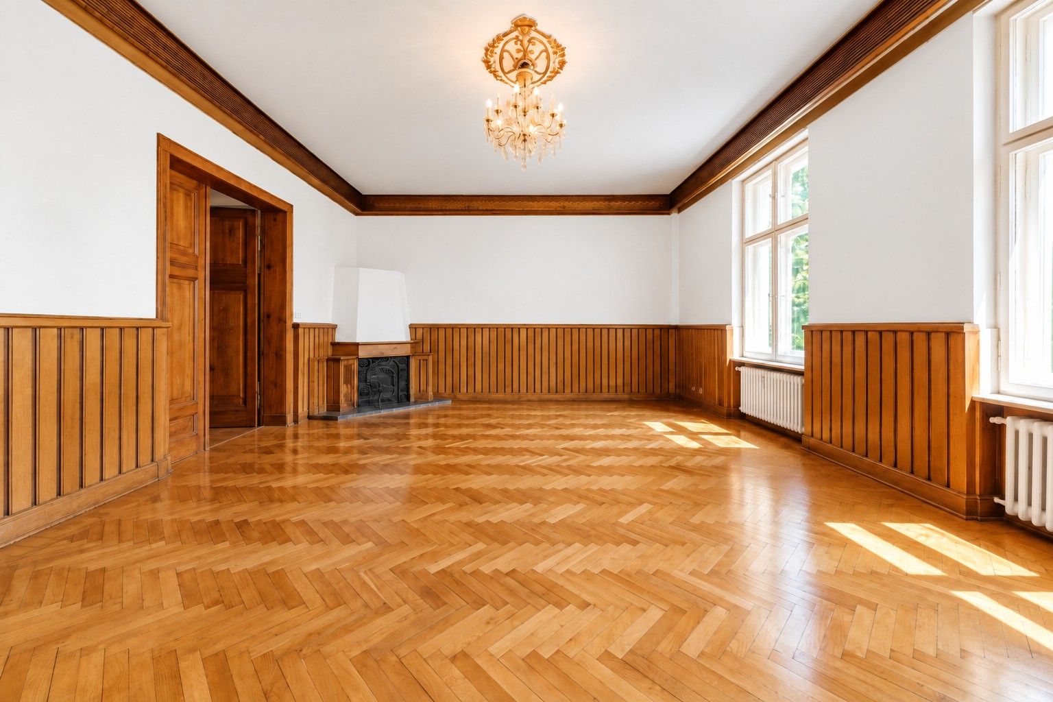 Leerer Raum mit Holzparkett, Holzverkleidung, Kamin, Kronleuchter und großen Fenstern.