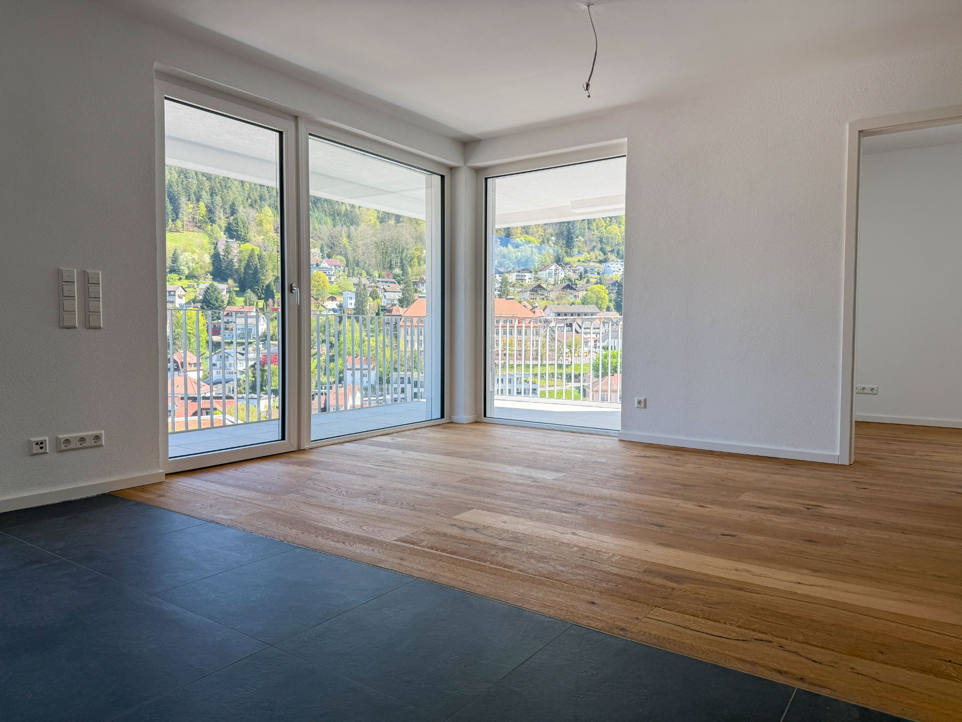 Helles Wohnzimmer mit Holzboden, großen Fenstern und Blick auf Balkon und Hügel.