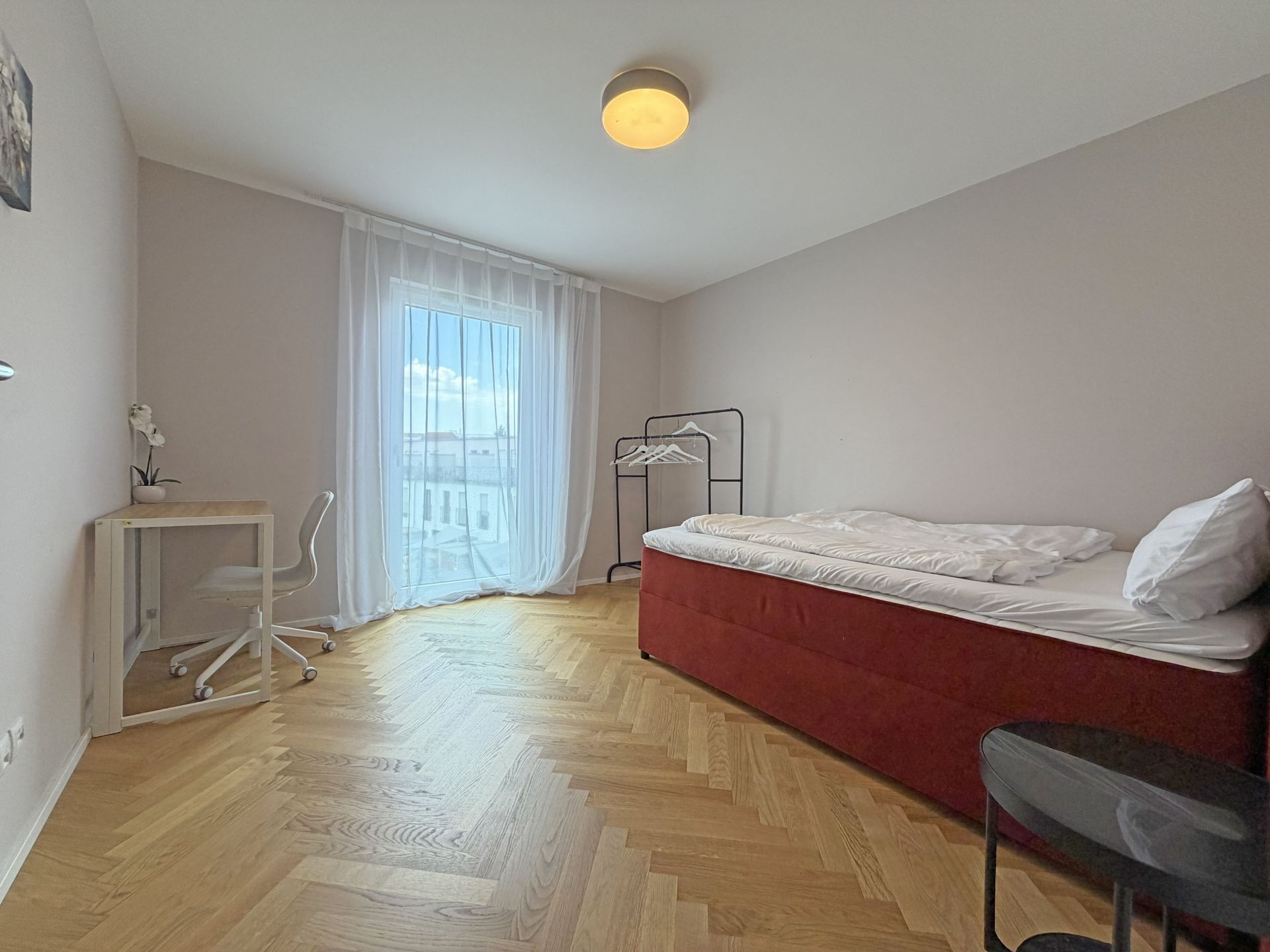 Helles Schlafzimmer mit Einzelbett, Schreibtisch, Stuhl und großem Fenster mit Vorhang.
