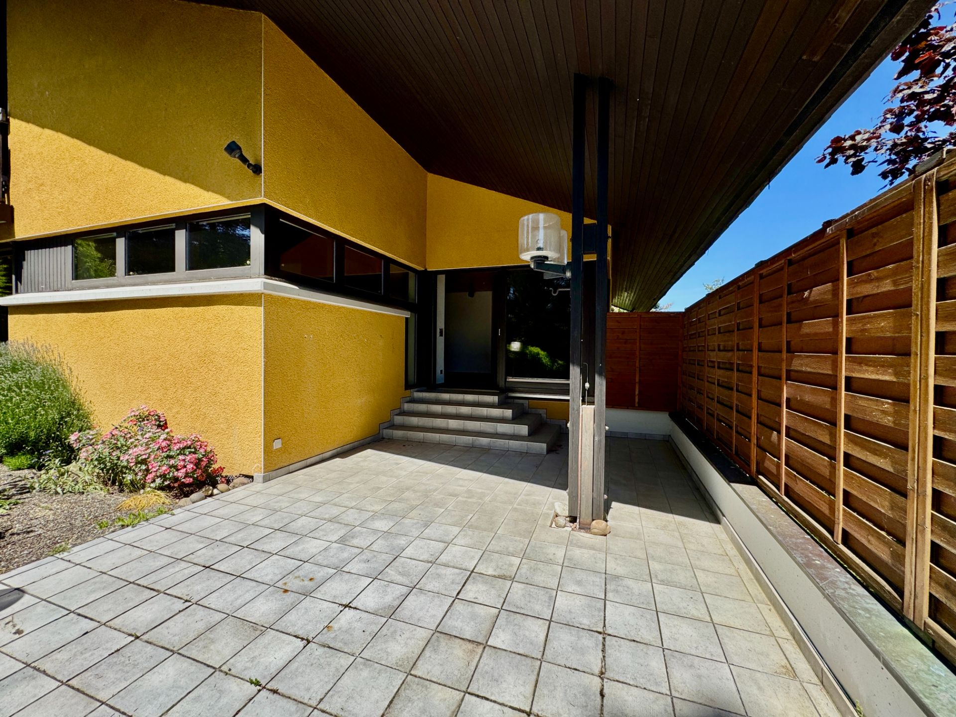Überdachte Terrasse mit gelber Wand, Fliesenboden und Stufen zum Hauseingang.