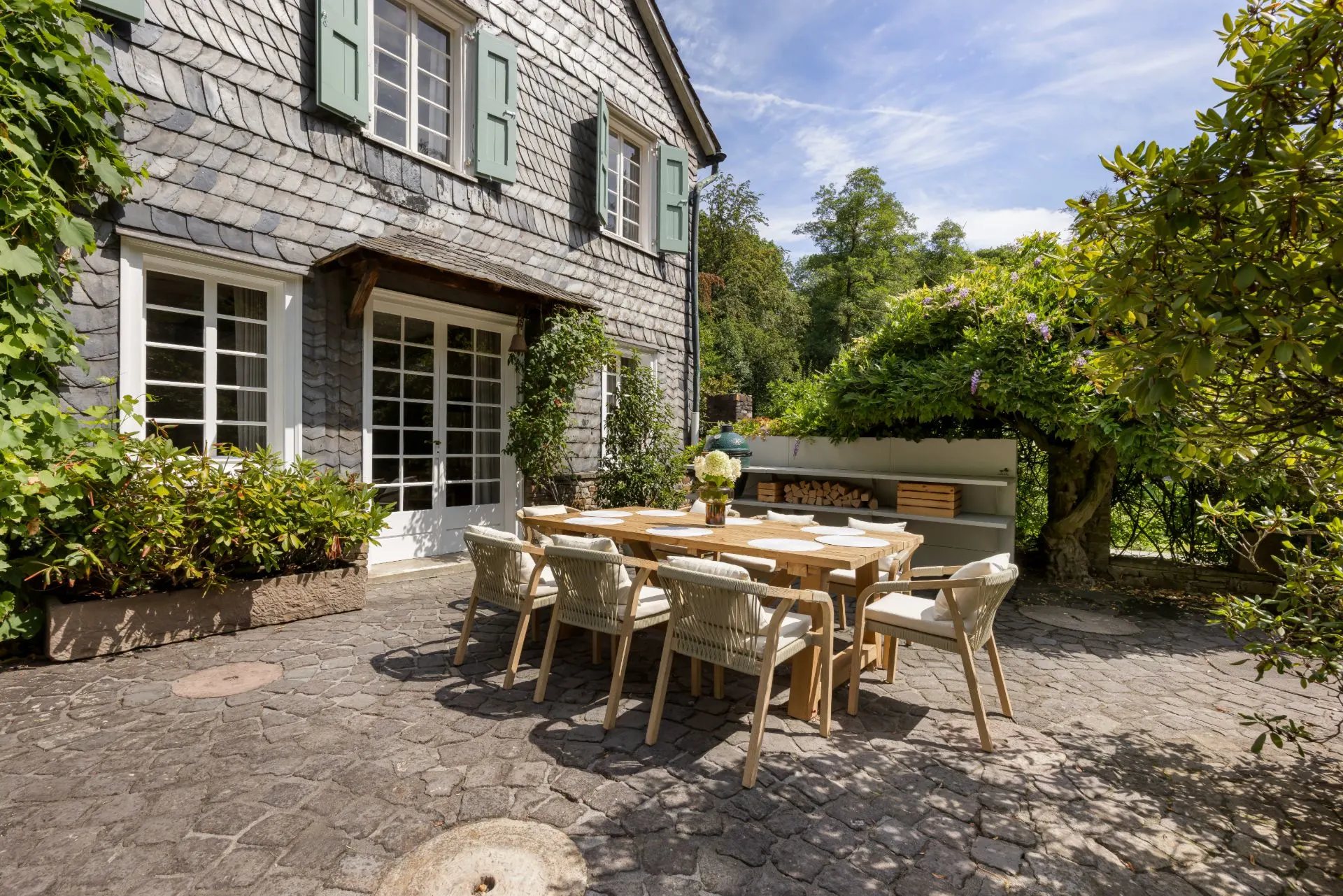 Gepflasterte Terrasse mit großem Holztisch und Stühlen vor einem Wohnhaus mit grünen Fensterläden.