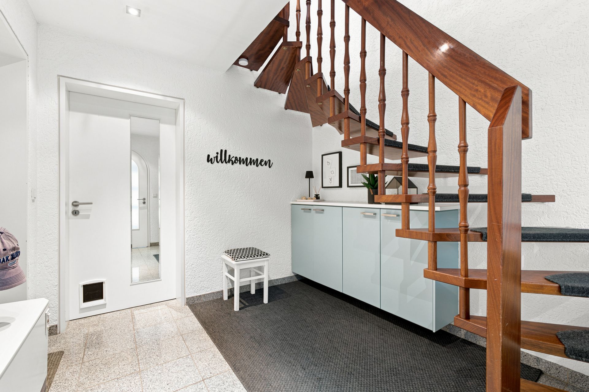 Heller Flur mit Holztreppe, Sideboard, Bank und Schriftzug 'willkommen' an der Wand.