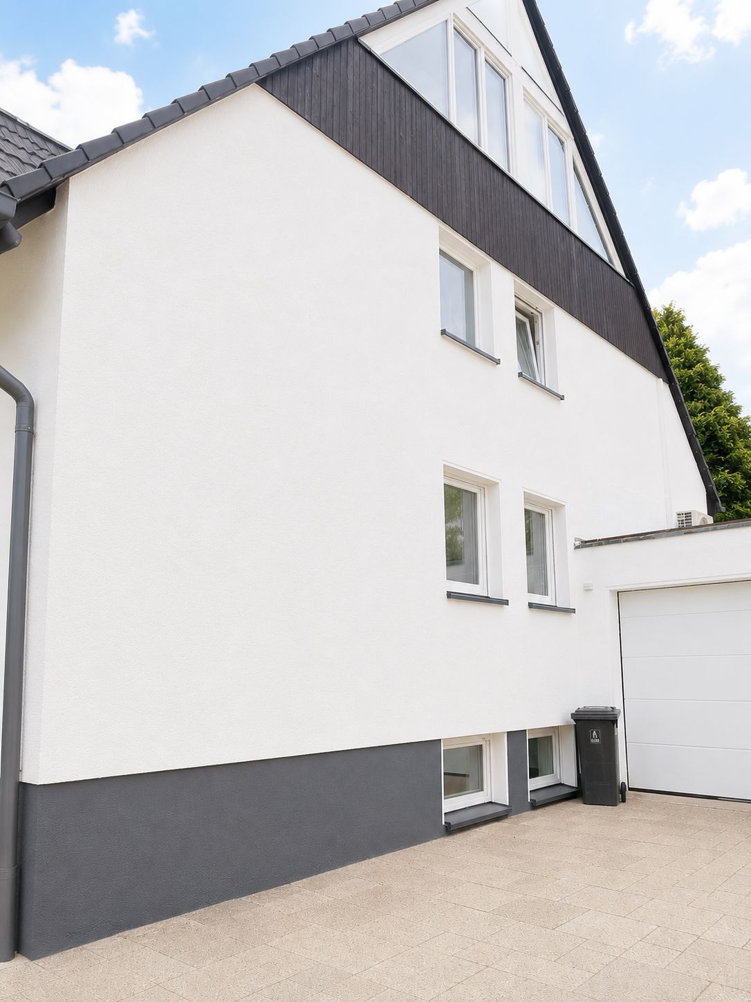 Moderne Hausfassade mit mehreren Fenstern und Garage, helle Mauer und dunkles Dach.