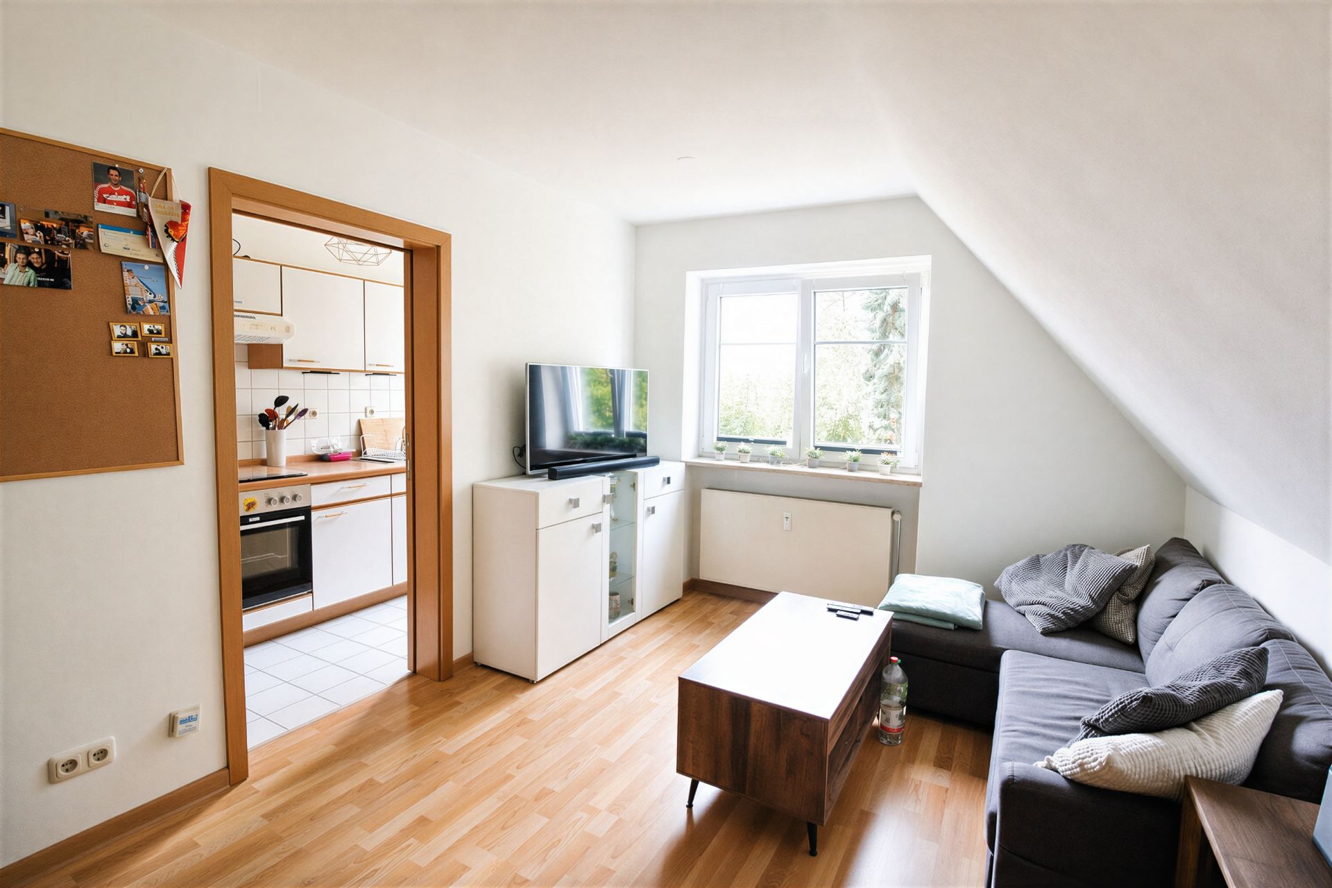 Wohnzimmer mit grauem Sofa, Holz-Couchtisch, Fernseher und Blick in eine helle Küche.