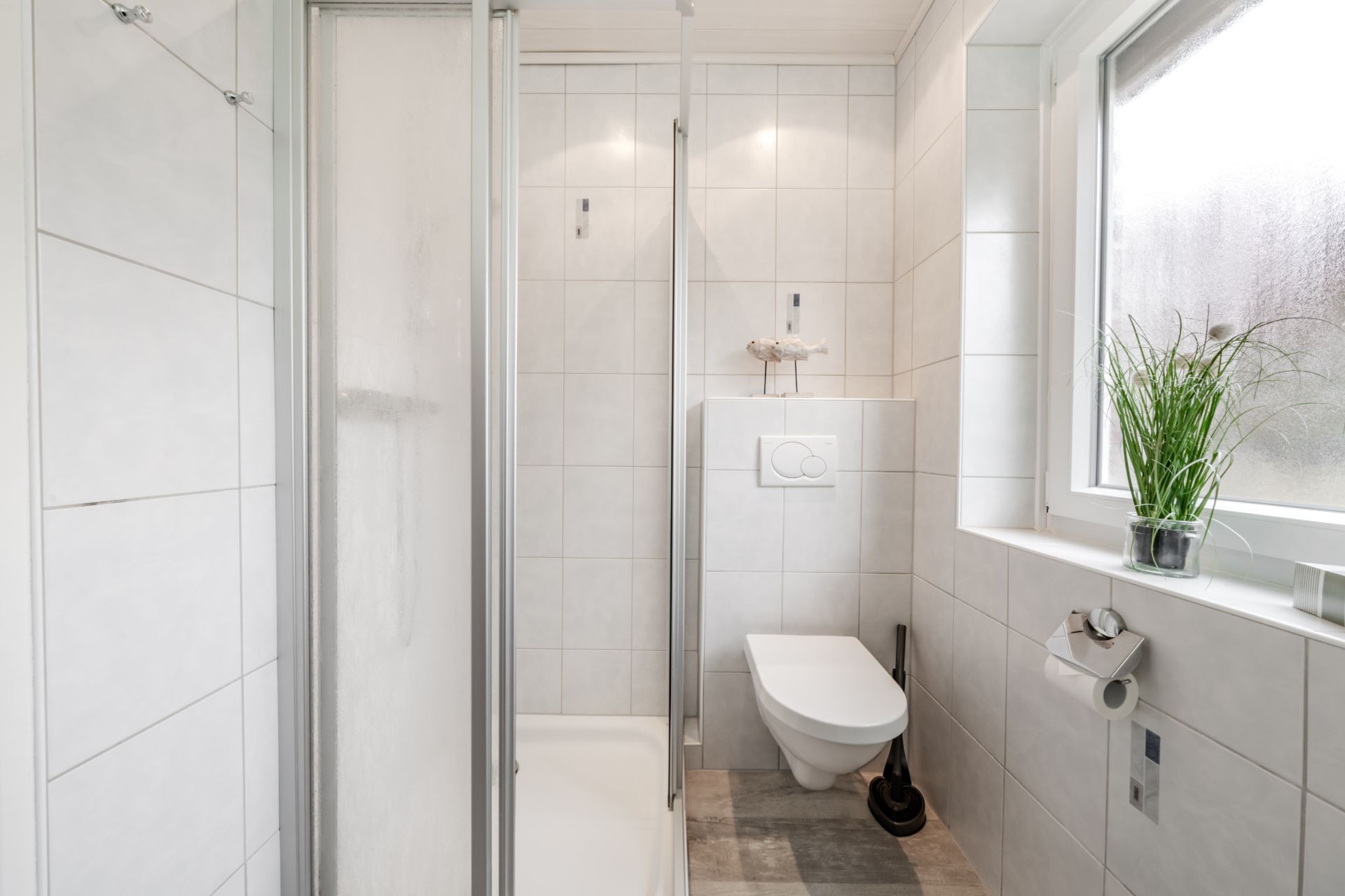 Modernes, weiß gefliestes Bad mit Dusche, Wand-WC, Fenster und Pflanze auf der Fensterbank.