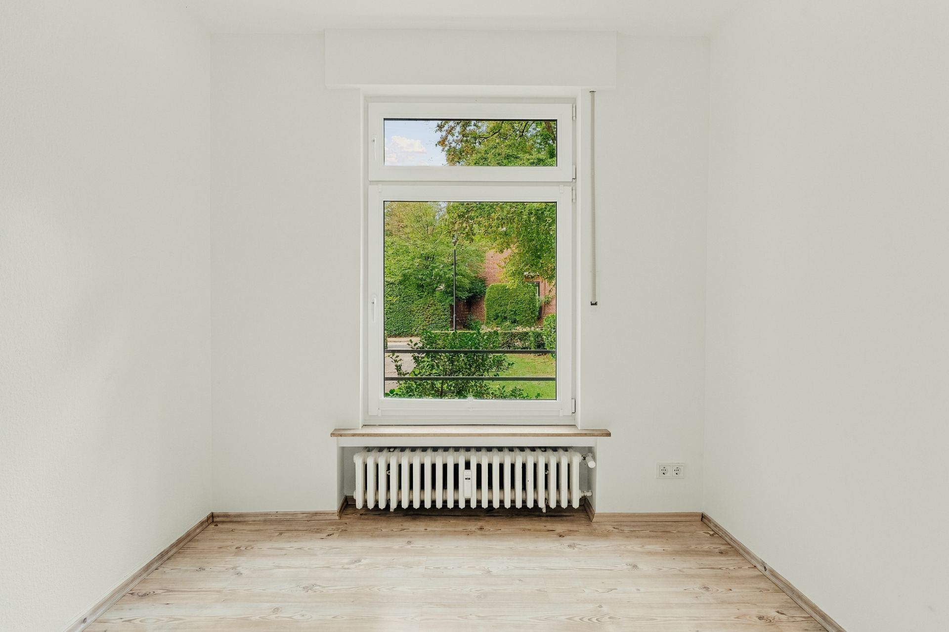 Heller, leerer Raum mit Holzboden, großem Fenster und Heizkörper darunter.