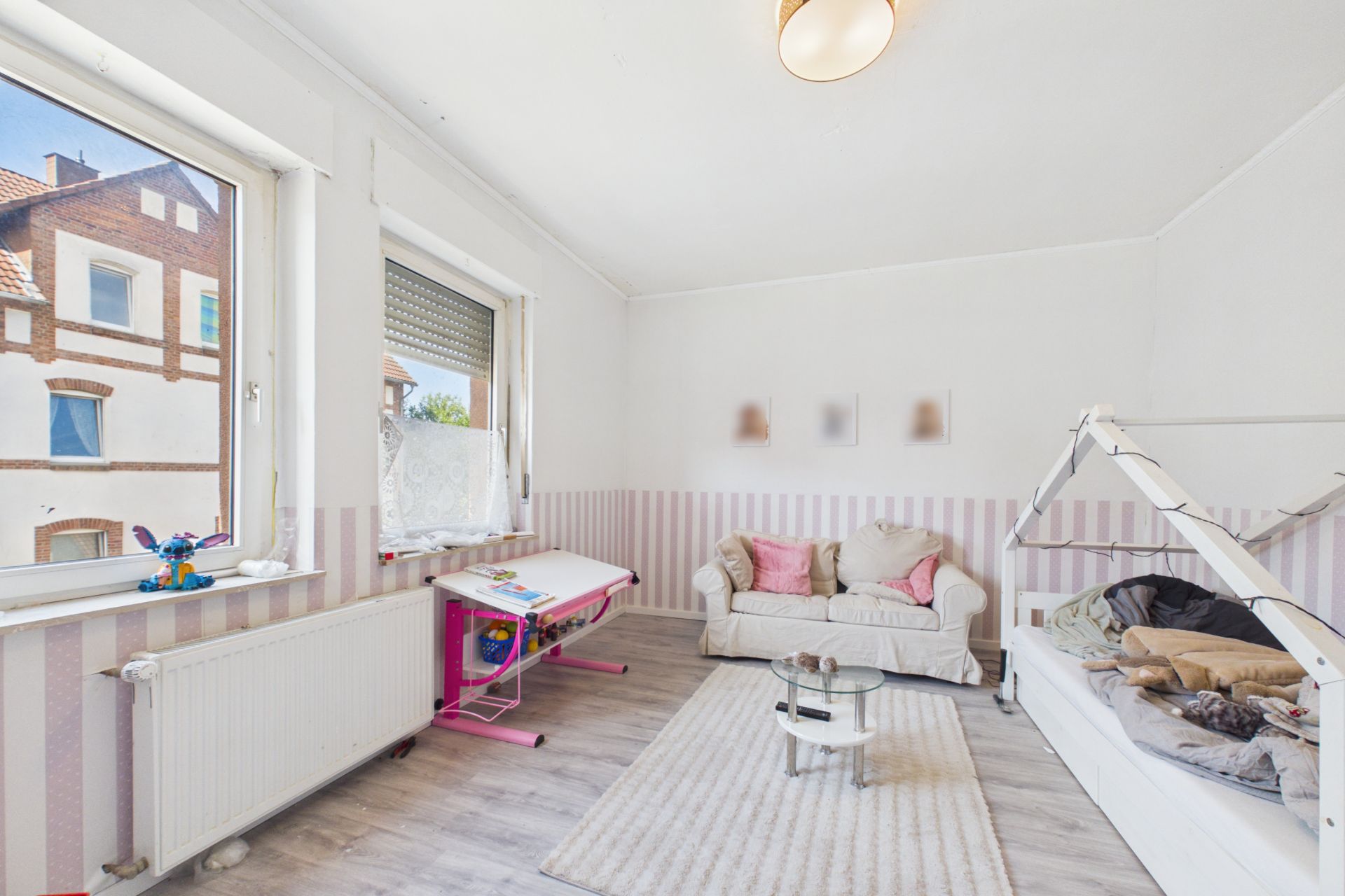 Helles Kinderzimmer mit zwei Fenstern, Sofa, kleinem Tisch, Schreibtisch und Kinderbett.