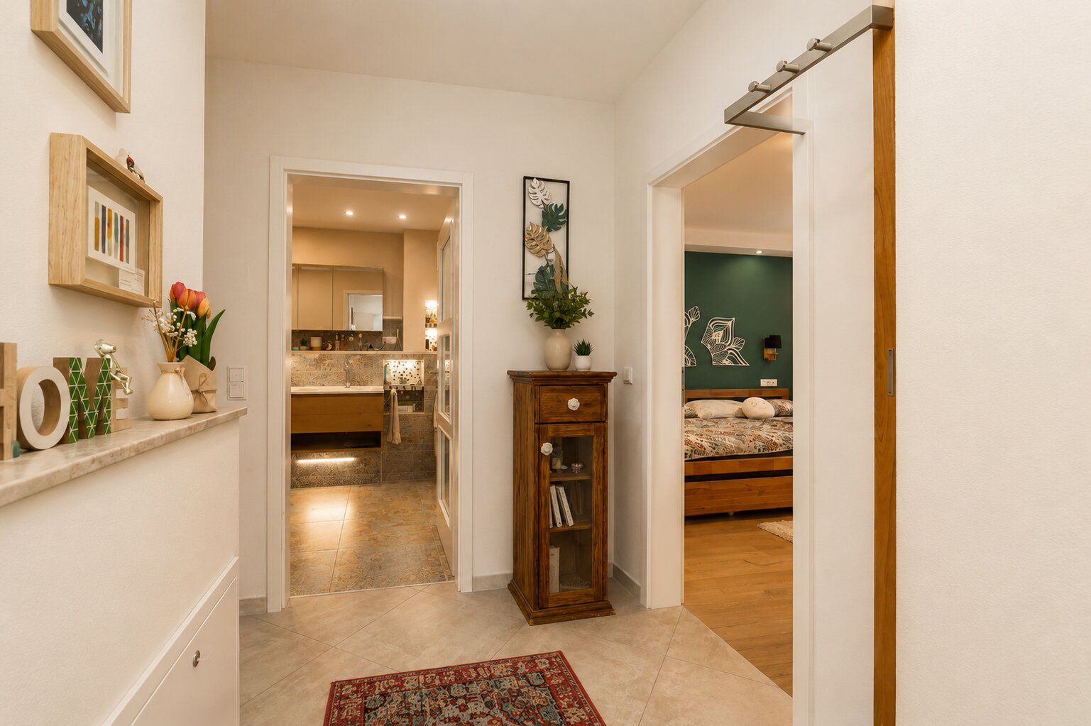 Heller Flur mit Teppich, Holzschrank, Wandbildern und Blick ins Schlafzimmer sowie Badezimmer.