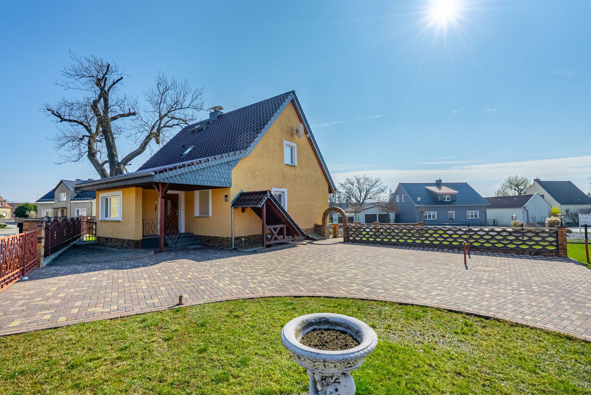 Gelbes Einfamilienhaus mit dunklem Dach, gepflasterter Hof und Rasen unter blauem Himmel.