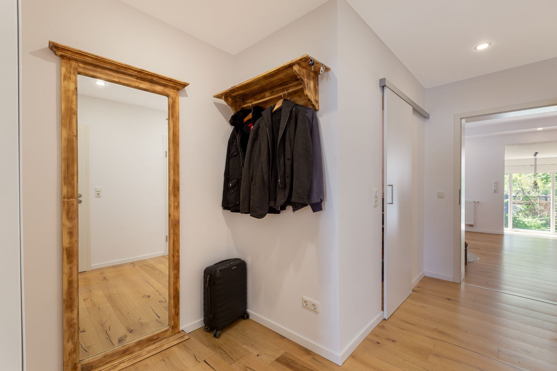 Flur mit Holzspiegel, Garderobe mit Jacken und einem kleinen schwarzen Koffer auf Holzboden.