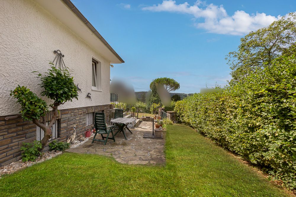 Steinterrasse mit Gartenmöbeln neben einem Haus und angrenzendem Rasen, umgeben von Hecken.