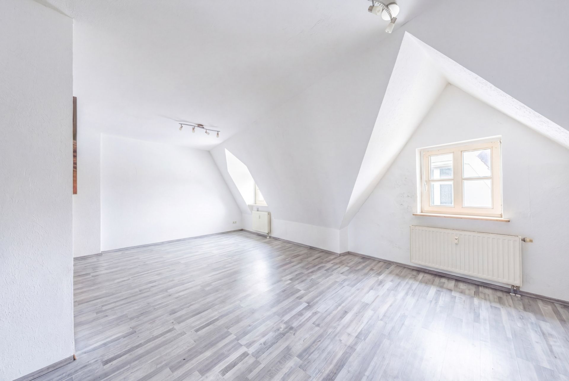 Heller, leerer Raum mit Dachschrägen, Holzboden und zwei Fenstern.
