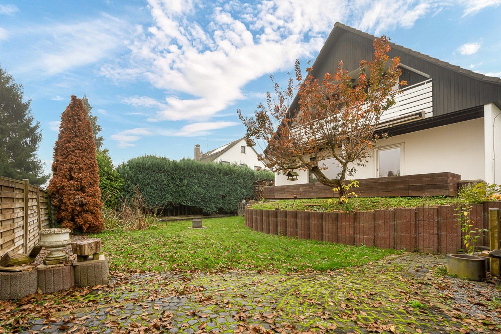 Garten mit Rasen, Bäumen, Steinterrasse und einem weißen Haus im Hintergrund.