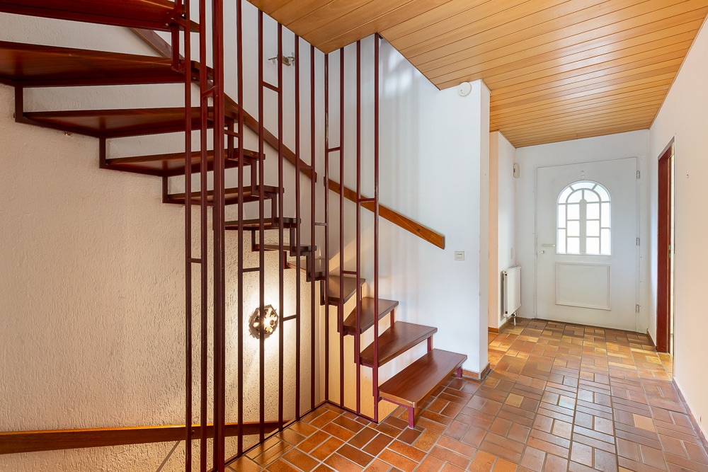 Flur mit gemauerter Treppe aus Holz und Metall, Fliesenboden und weißer Eingangstür.