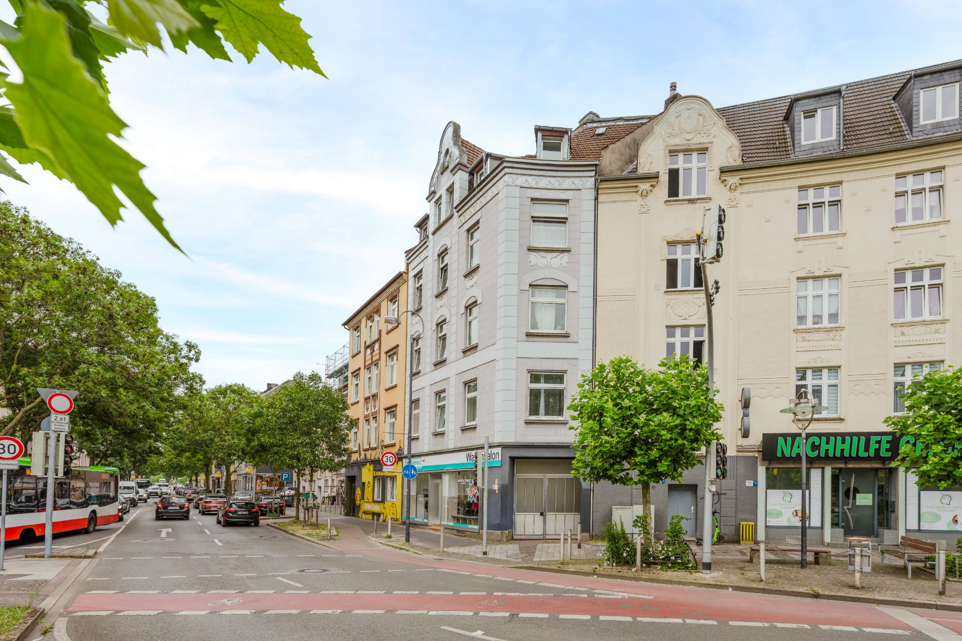 Wohnhäuser an einer Straße mit Bäumen, Geschäften im Erdgeschoss und vorbeifahrenden Autos.