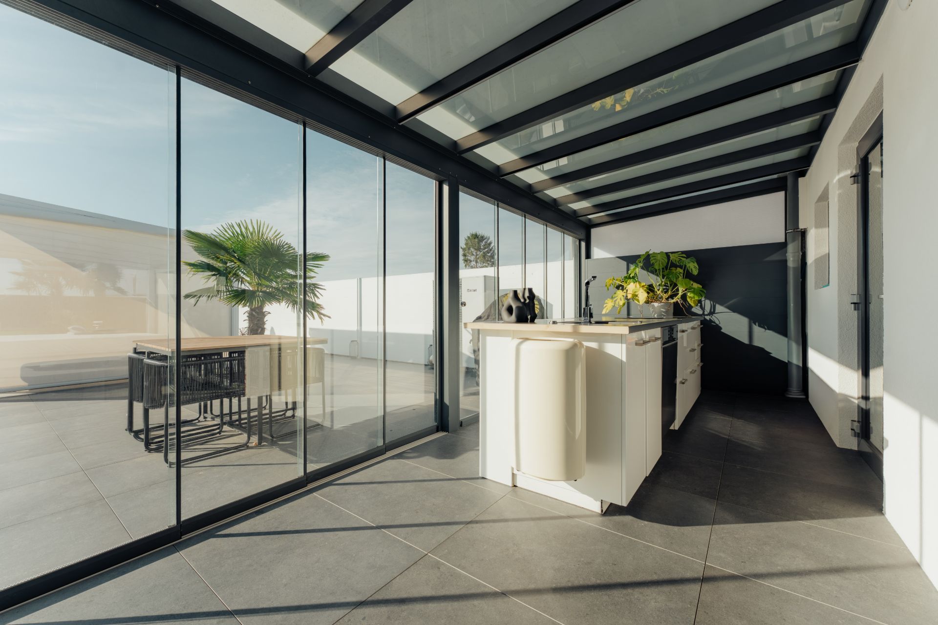Moderne verglaste Terrasse mit Kücheninsel und Blick auf eine Palme draußen.