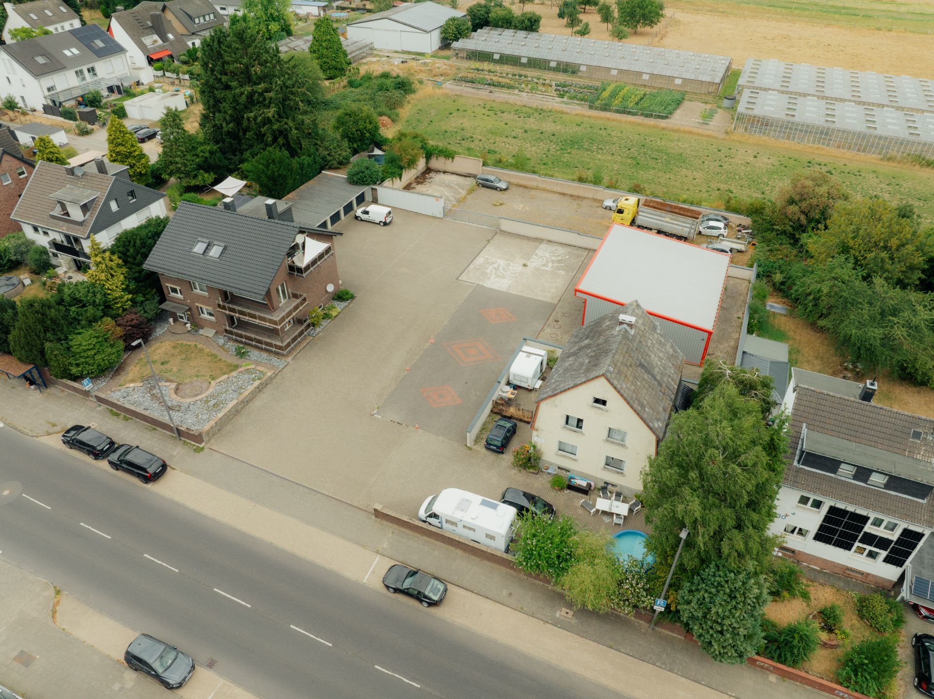 Luftaufnahme eines Wohnhauses mit großem Hof, mehreren parkenden Autos und angrenzender Straße.