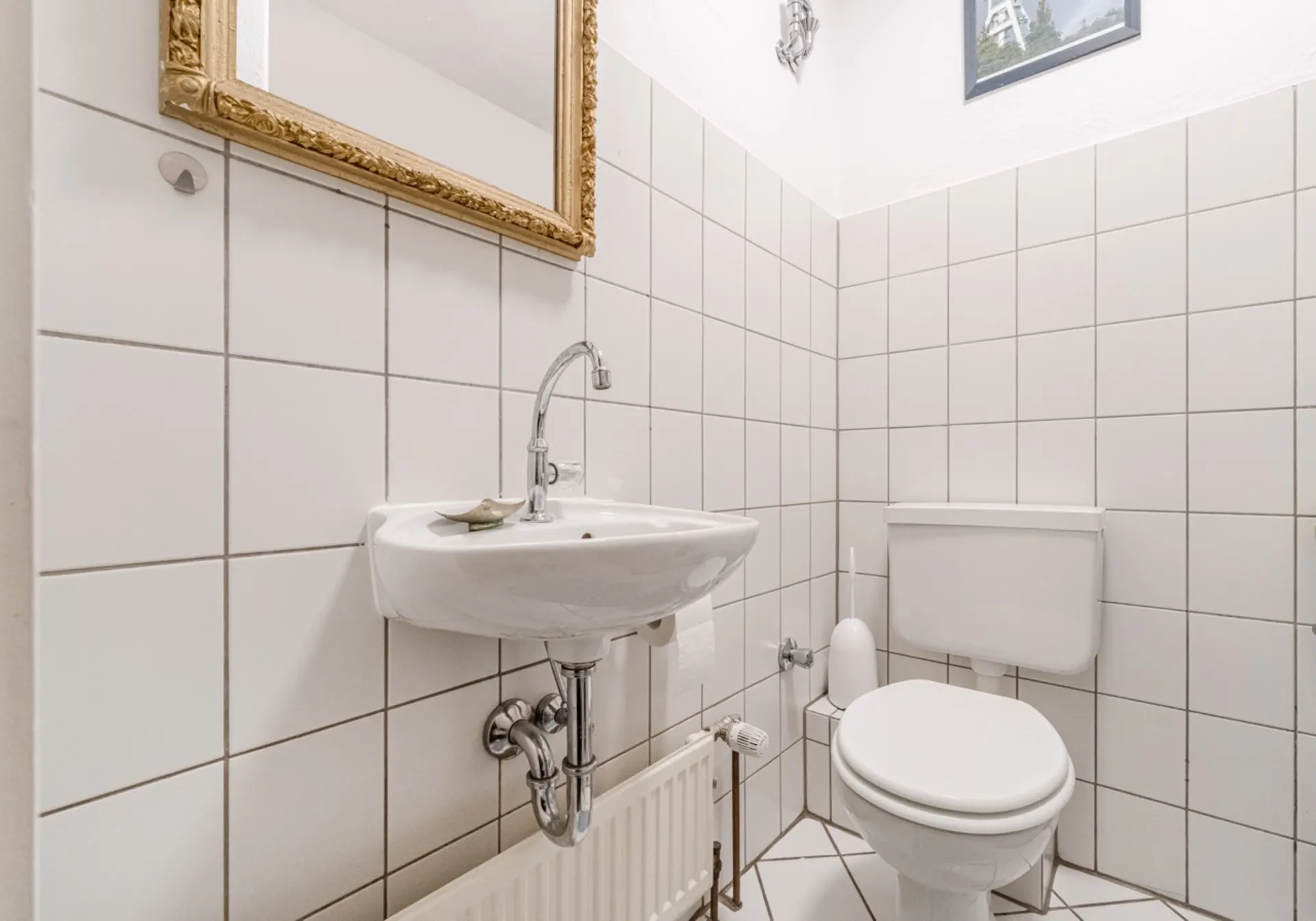 Kleines Badezimmer mit weißem Waschbecken, Toilette und Spiegel an der gefliesten Wand.