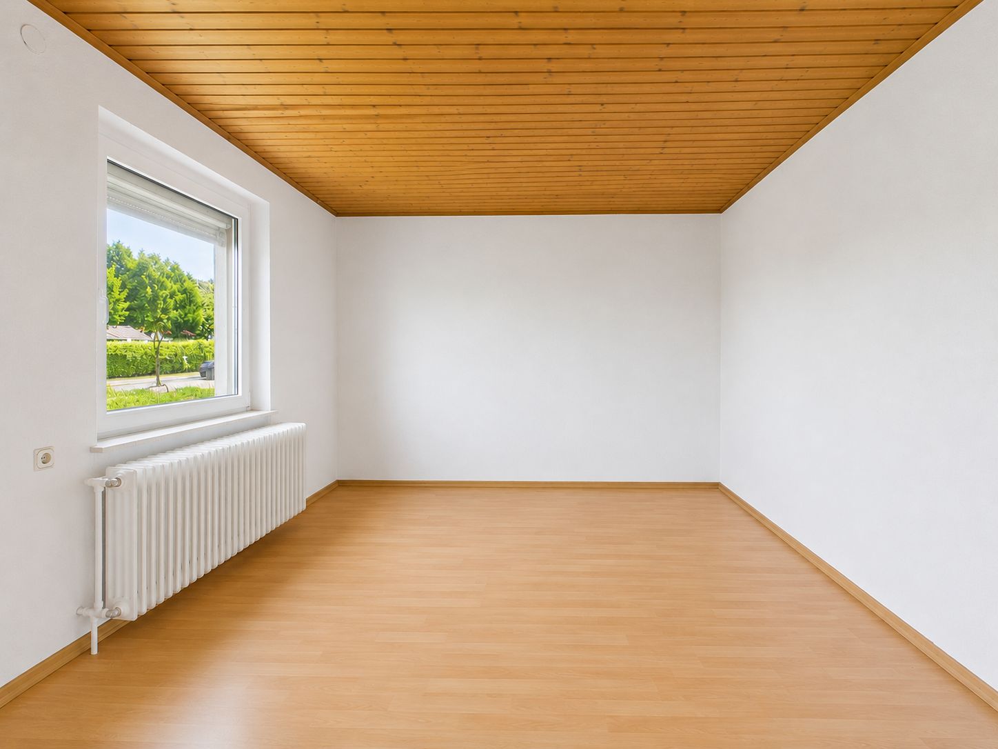 Leerer Raum mit Holzdecke, weißem Heizkörper und großem Fenster mit Blick ins Grüne.