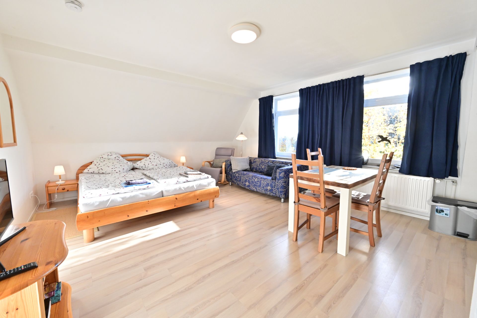 Helles Zimmer mit Doppelbett, Esstisch, Sofa und zwei großen Fenstern mit dunklen Vorhängen.
