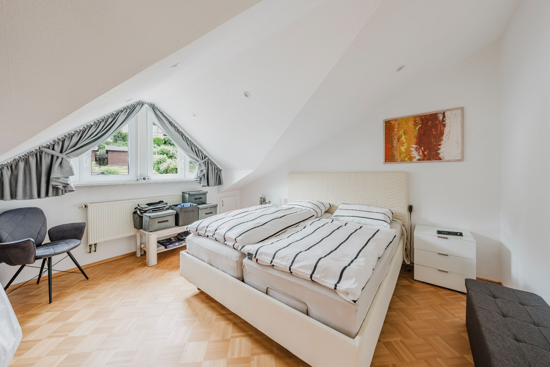 Schlafzimmer mit Doppelbett unter Dachschräge, Fenster mit grauen Vorhängen, Stuhl und Kommode.