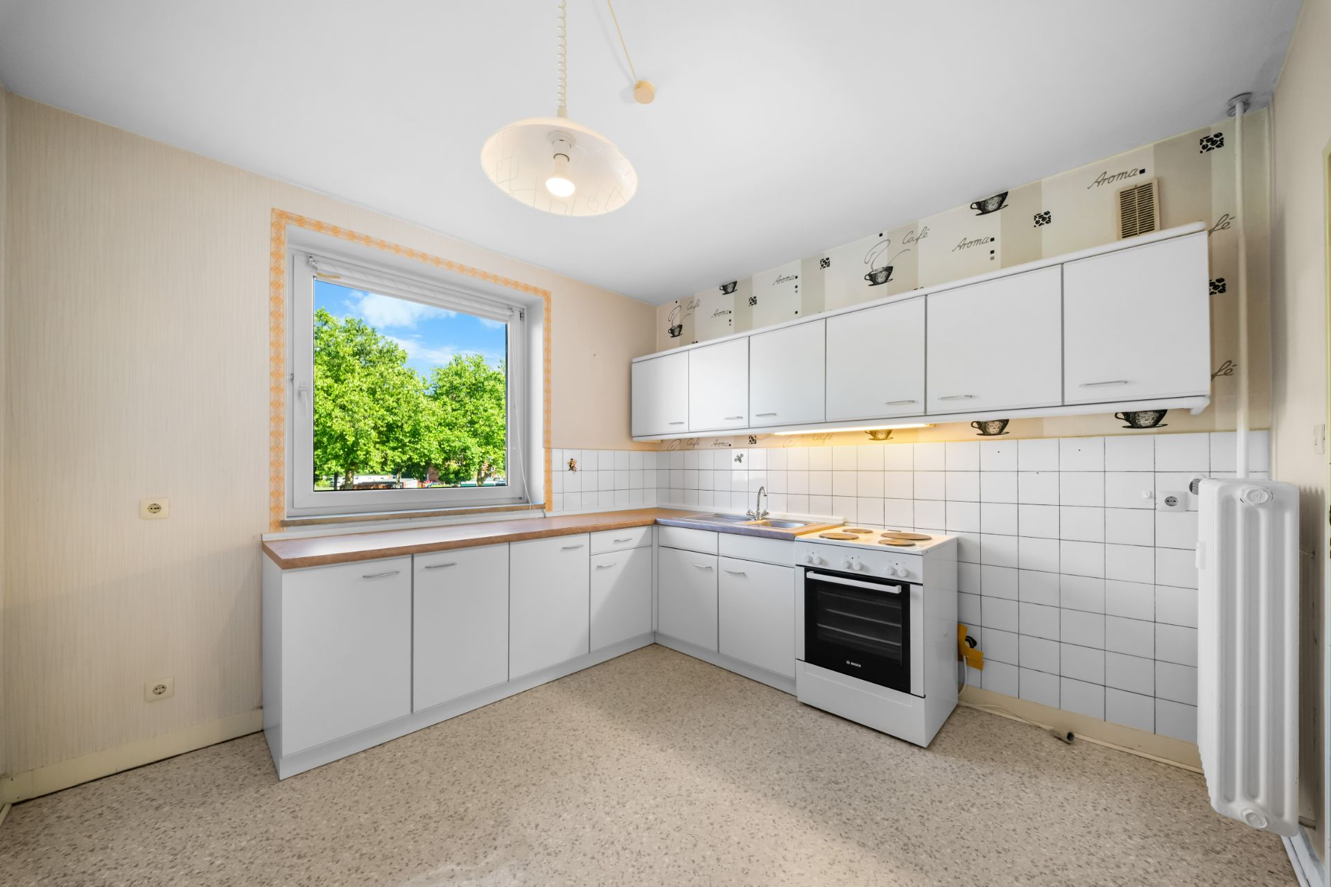 Helle Einbauküche mit weißen Schränken, Herd, Spüle und Fenster mit Blick ins Grüne.