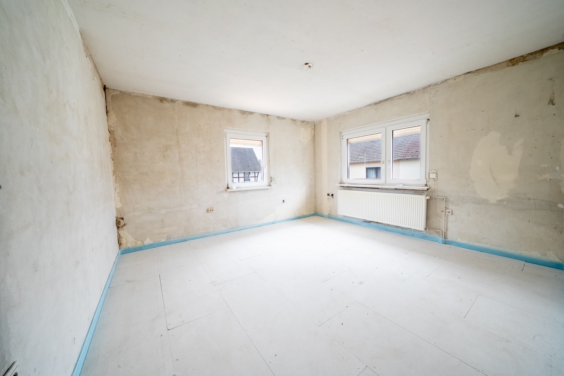 Leerer, renovierungsbedürftiger Raum mit zwei Fenstern und sichtbarem Heizkörper an der Wand.