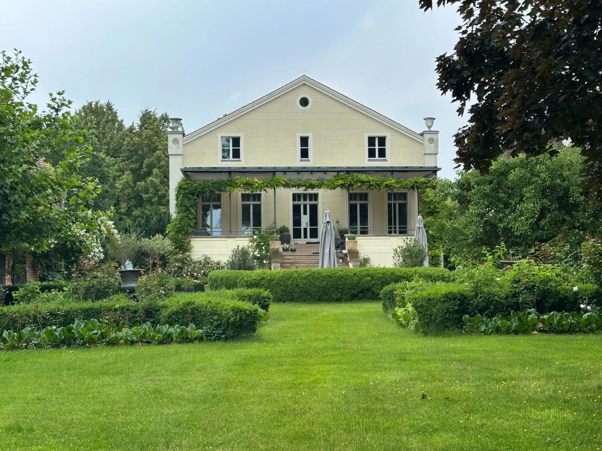 Großes gelbes Haus mit Terrasse und grünem Garten davor, umgeben von Bäumen und Pflanzen.