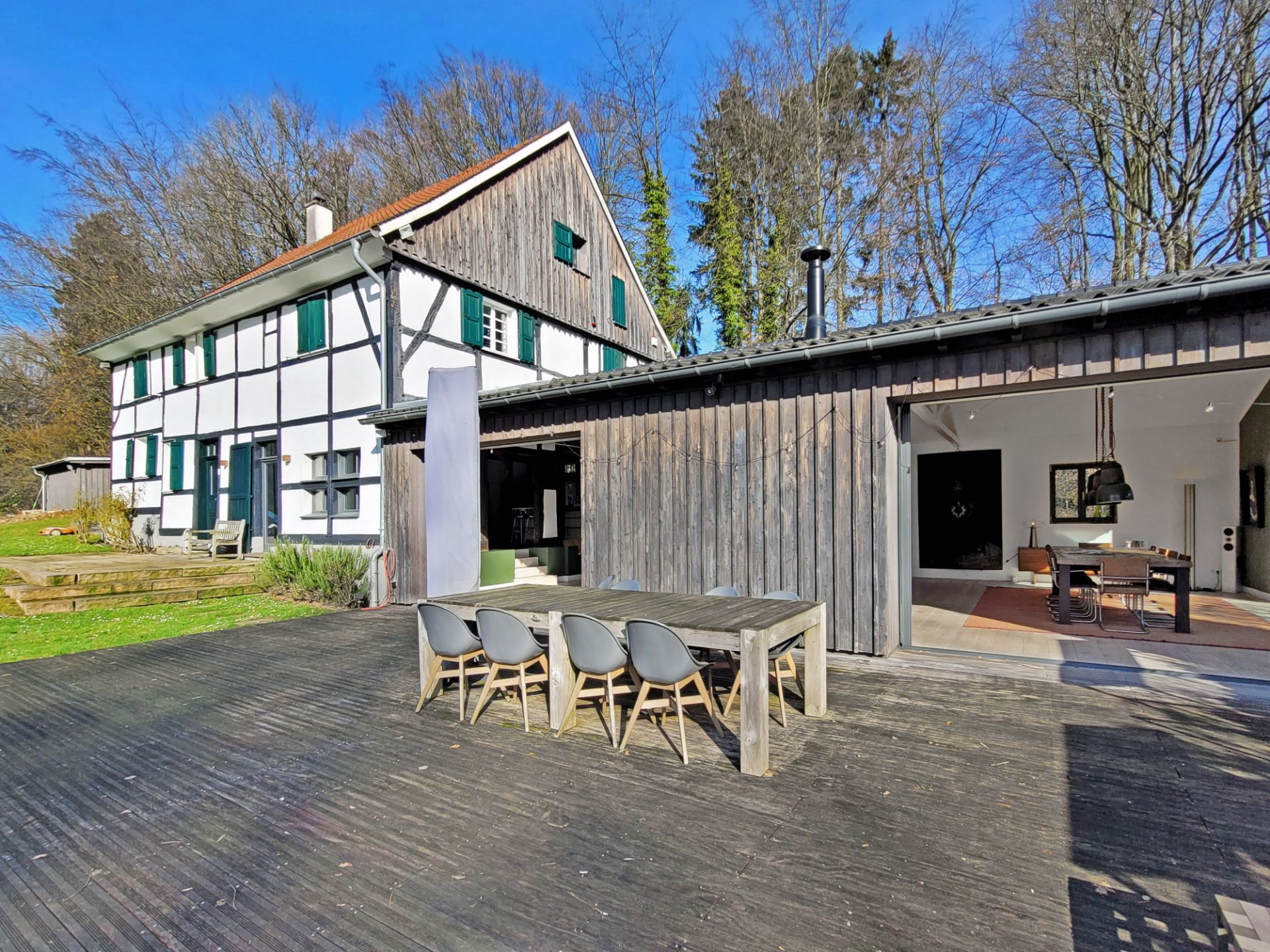Wohnhaus mit großer Terrasse, Holztisch und sechs Stühlen im Garten, umgeben von Bäumen.