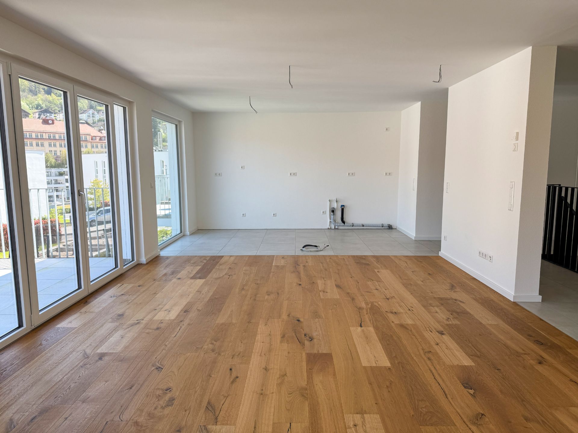 Helles Wohnzimmer mit Parkettboden, großen Fenstern und einem Bereich mit Fliesen für eine Küche.