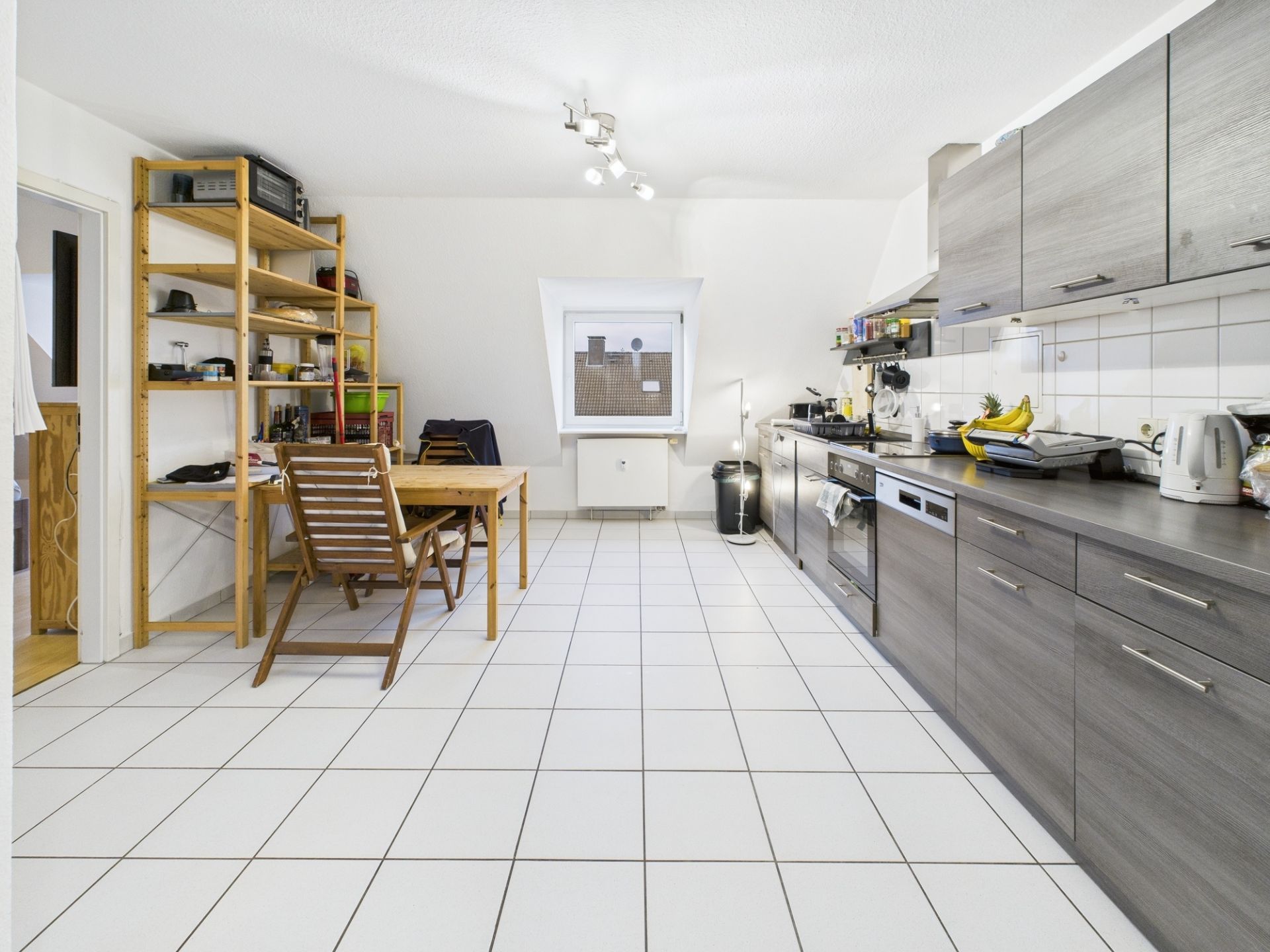 Moderne Küche mit grauen Schränken, Esstisch und Holzregal auf weiß gefliestem Boden.