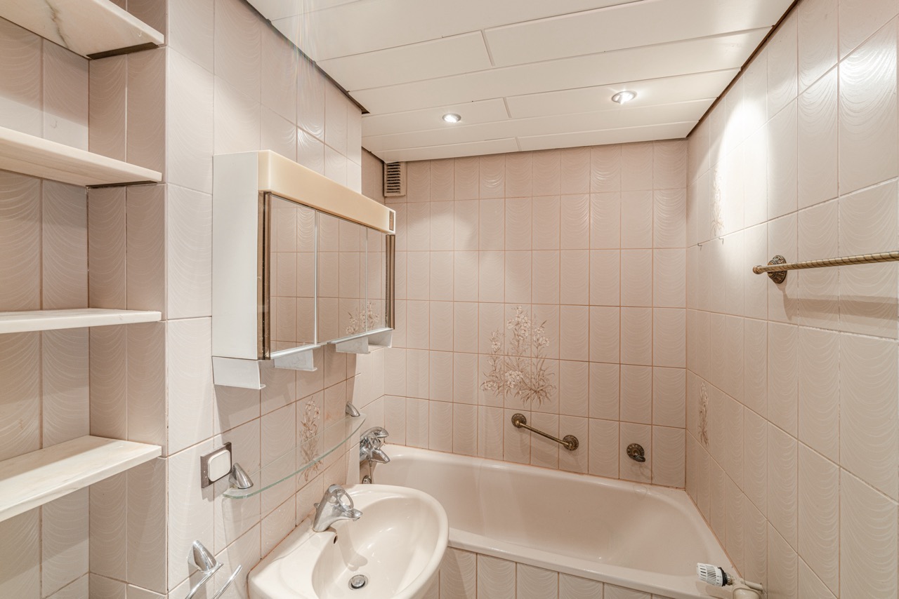 Kleines Badezimmer mit beiger Badewanne, Waschbecken, Wandspiegel und hellen Fliesen mit Blumenmuster.