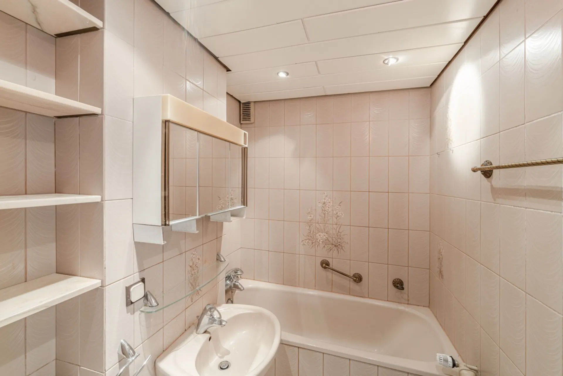Kleines Badezimmer mit beiger Badewanne, Waschbecken, Wandspiegel und hellen Fliesen mit Blumenmuster.