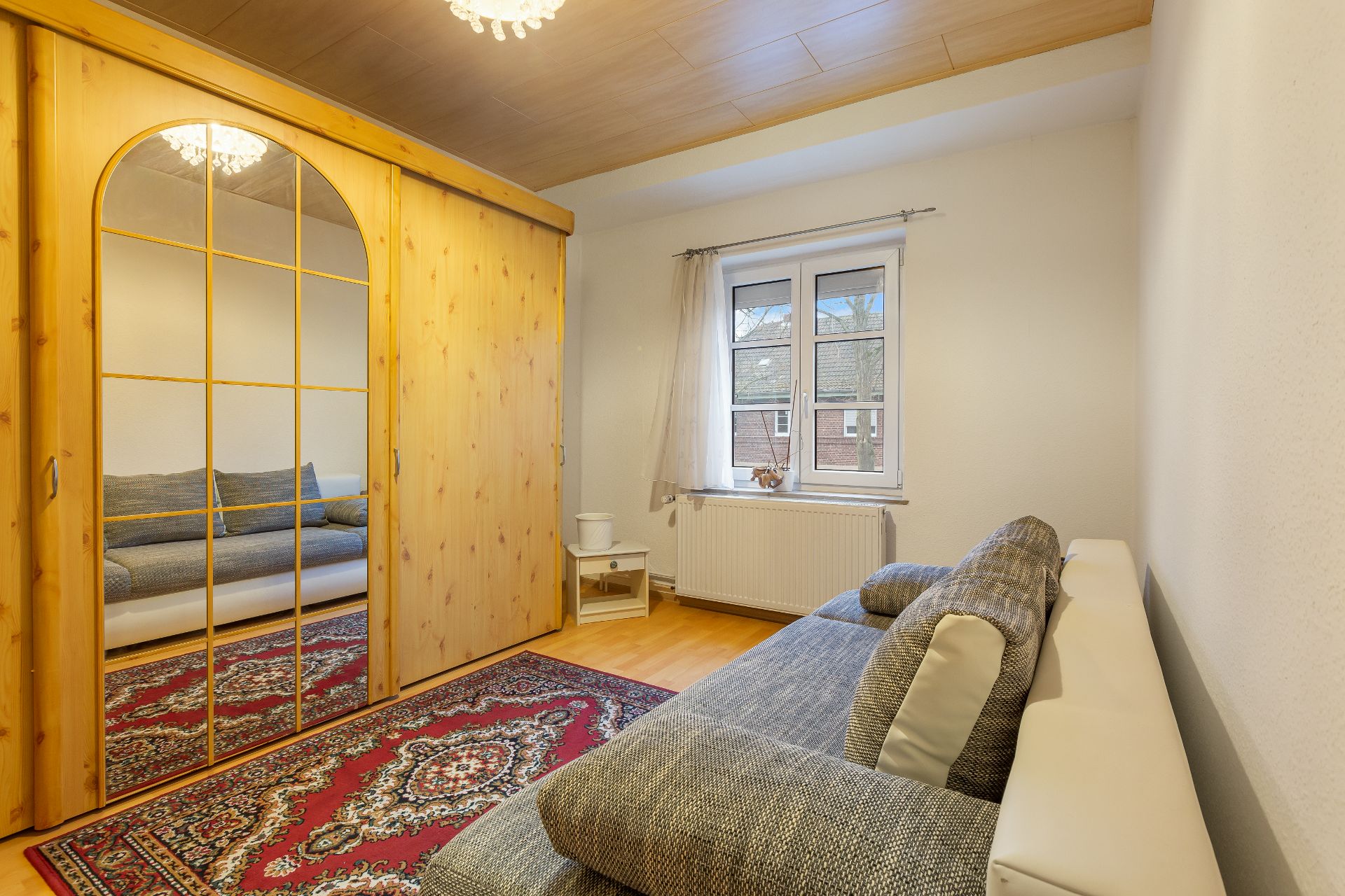 Zimmer mit grauem Sofa, großem Holzkleiderschrank mit Spiegel, Teppich und Fenster mit Blick nach draußen.