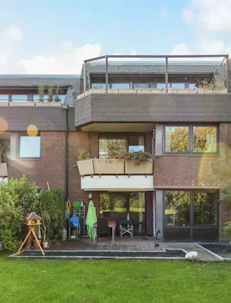 Rückseite eines Mehrfamilienhauses mit Balkonen, Terrasse und grüner Wiese im Vordergrund.