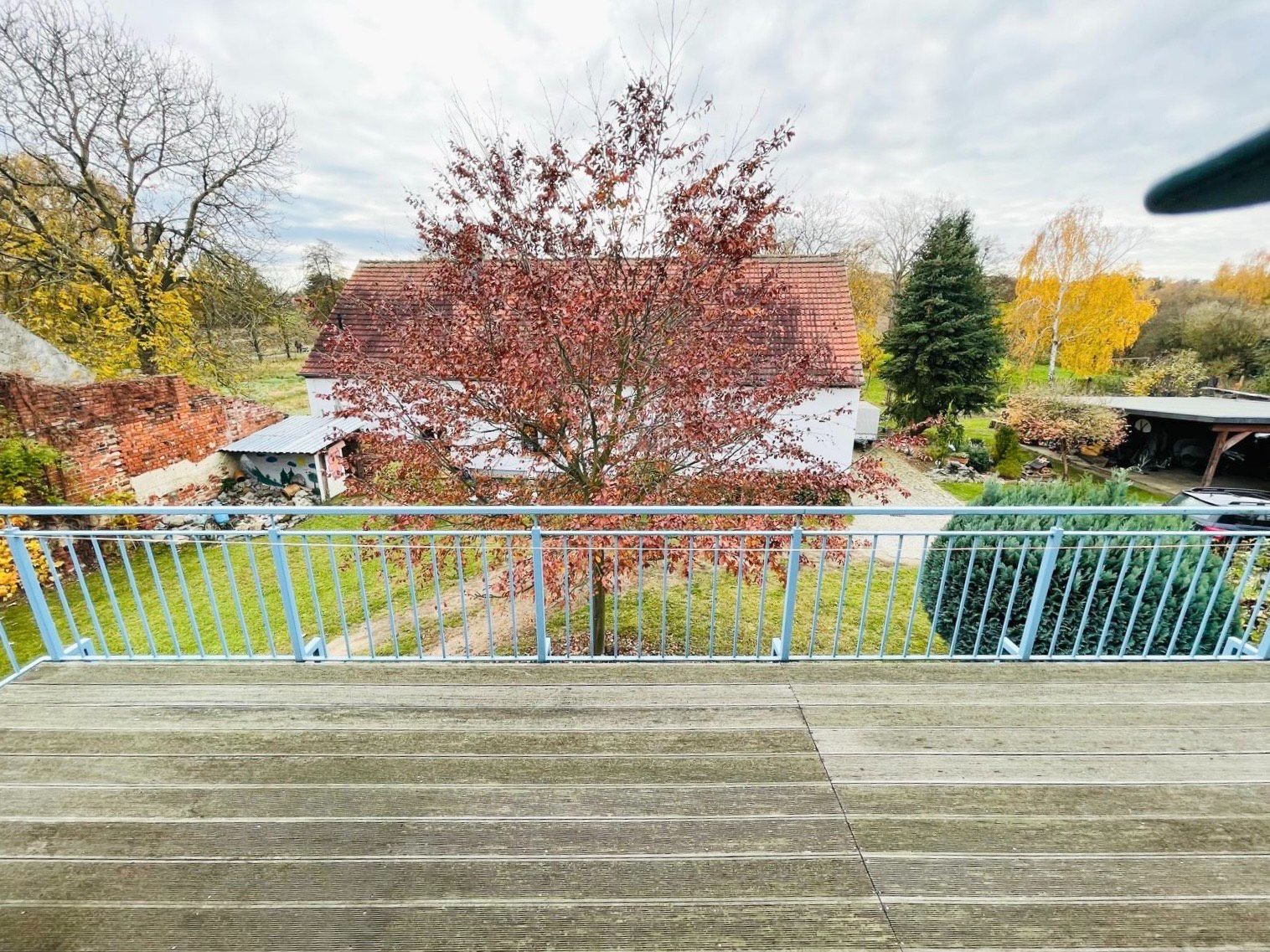 Blick von einem Balkon auf einen Garten mit Herbstbäumen und Haus im Hintergrund.