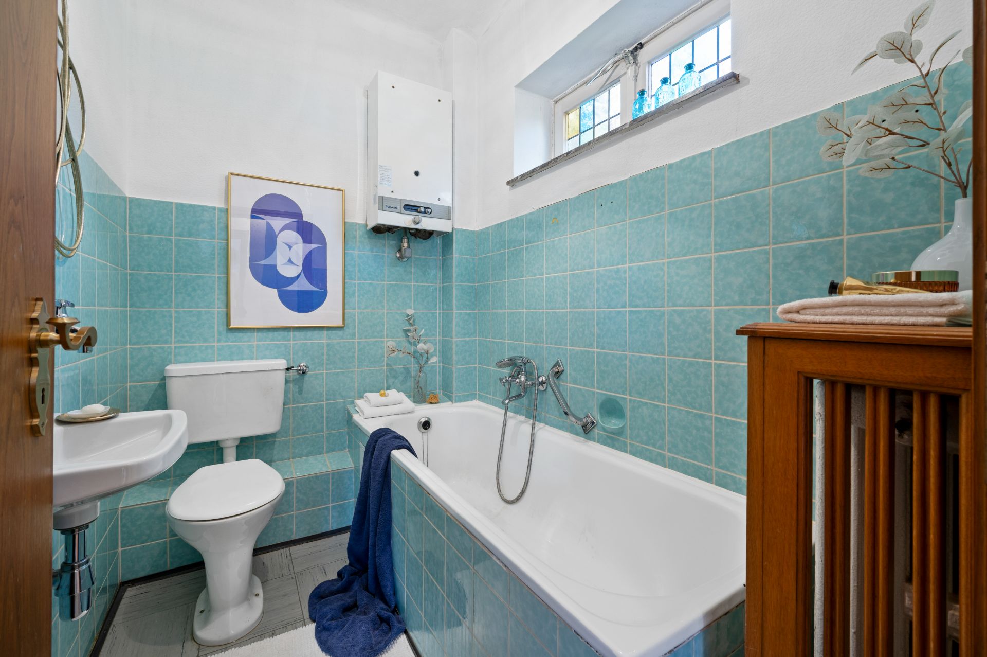 Kleines Badezimmer mit Badewanne, WC, Waschbecken und türkisfarbenen Fliesen.