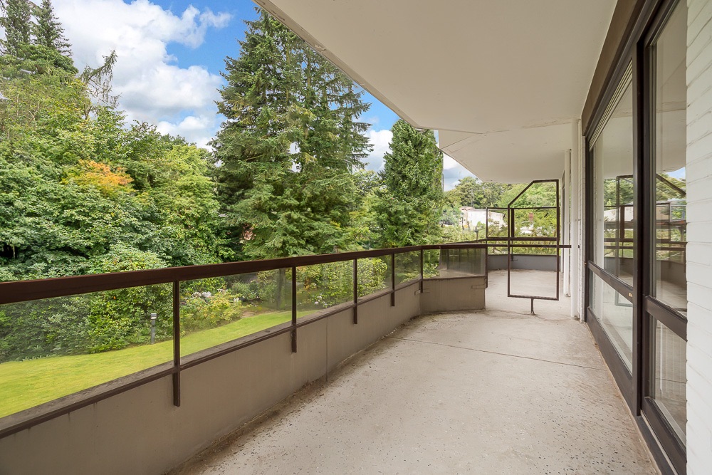 Breiter Balkon mit Glasgeländer und Blick auf einen grünen Garten mit vielen Bäumen.