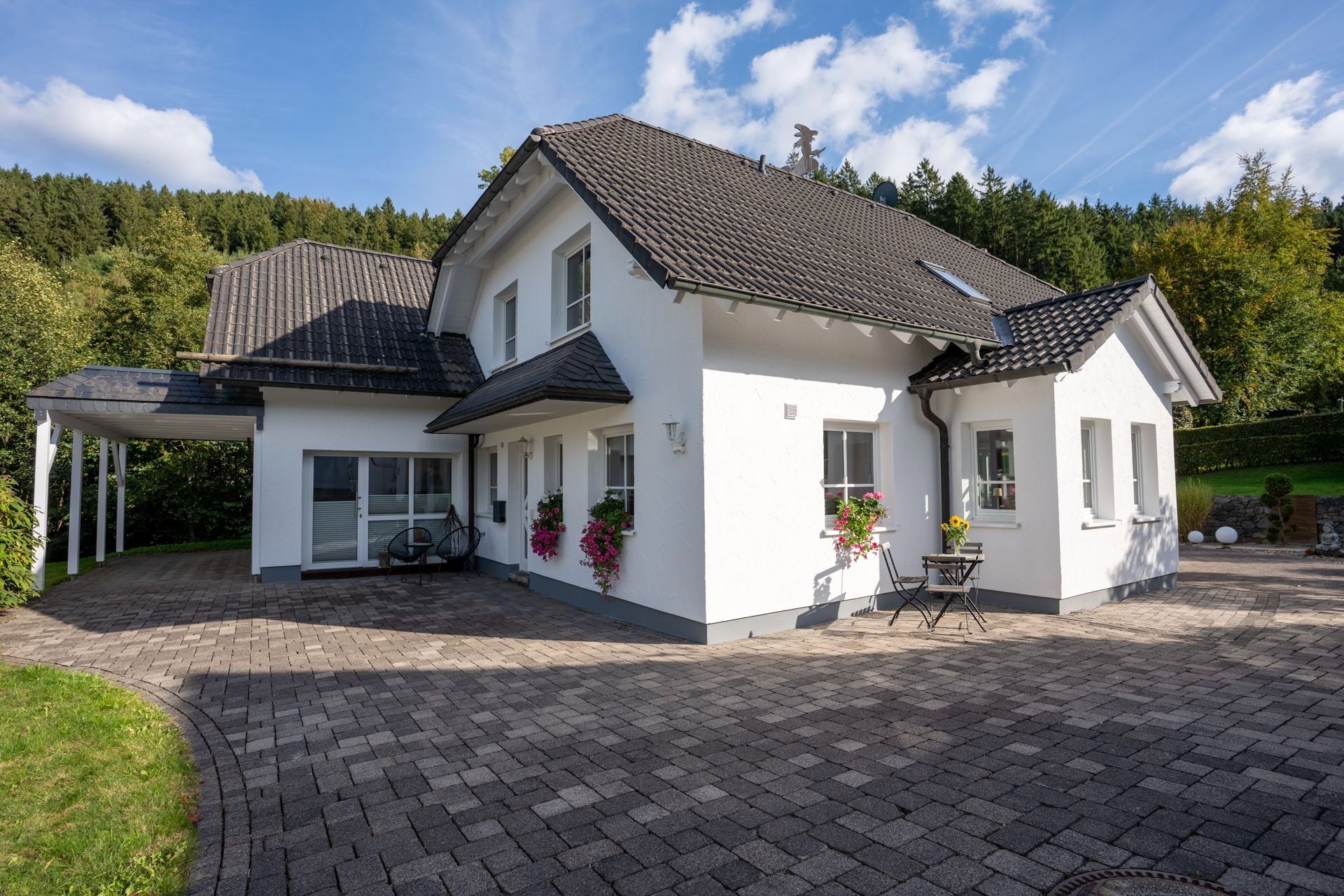 Modernes weißes Einfamilienhaus mit dunklem Dach, gepflastertem Hof und Garten im Hintergrund.