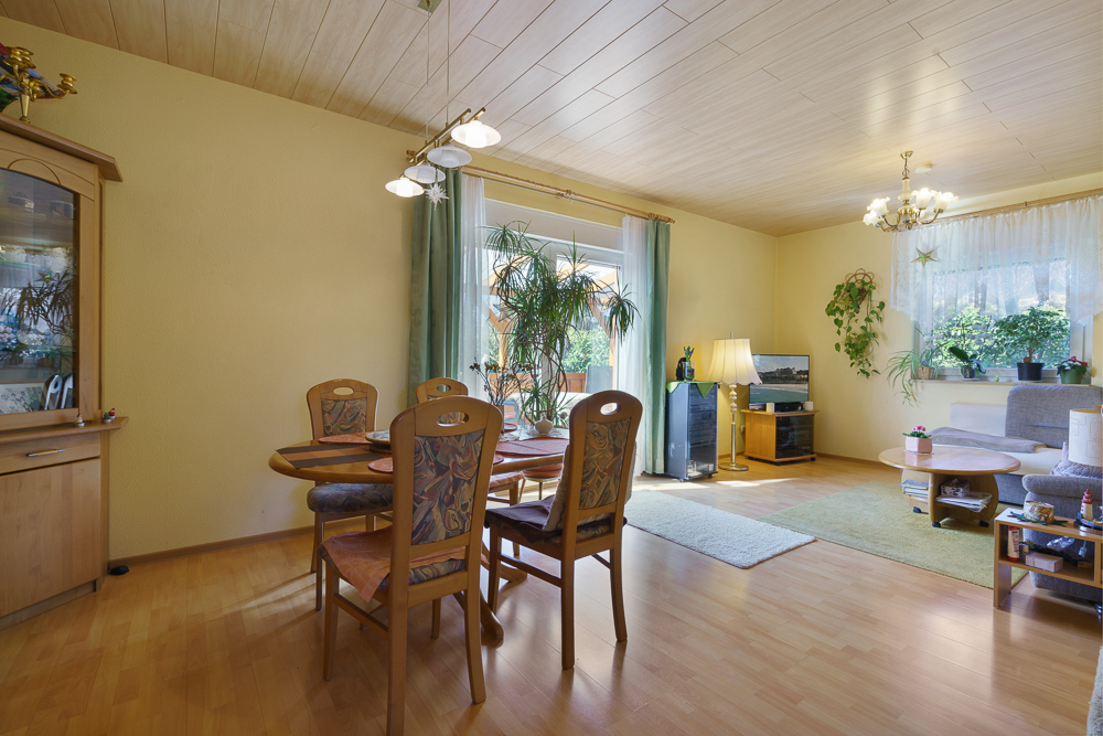 Wohnzimmer mit Essbereich, Holzmöbeln, Pflanzen, Teppichen und großen Fenstern mit Vorhängen.
