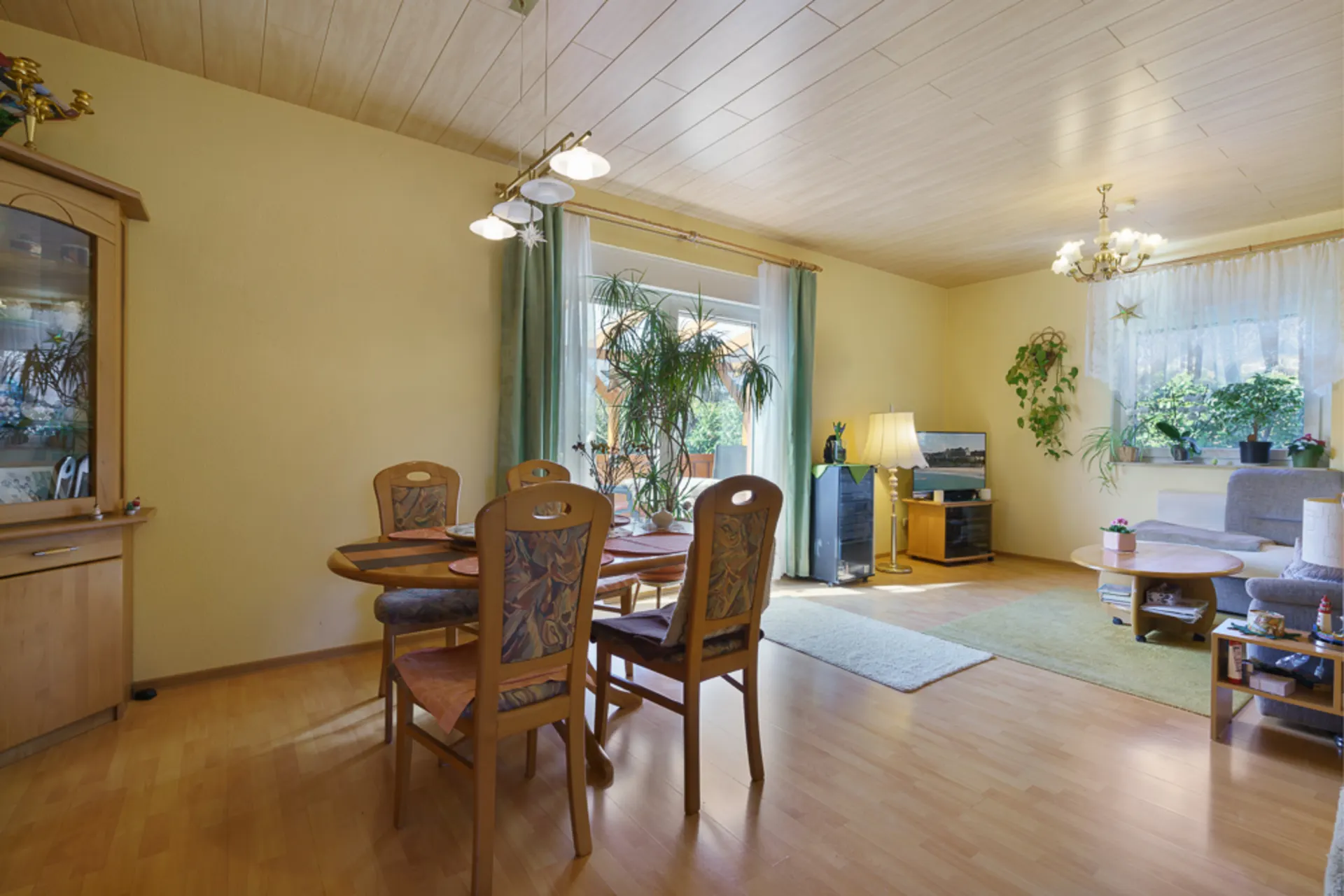 Wohnzimmer mit Essbereich, Holzmöbeln, Pflanzen, Teppichen und großen Fenstern mit Vorhängen.