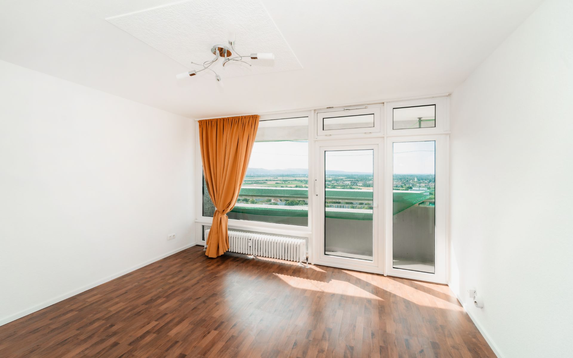 Helles Wohnzimmer mit Holzboden, großem Fenster, Glastür zum Balkon und orangefarbener Gardine.