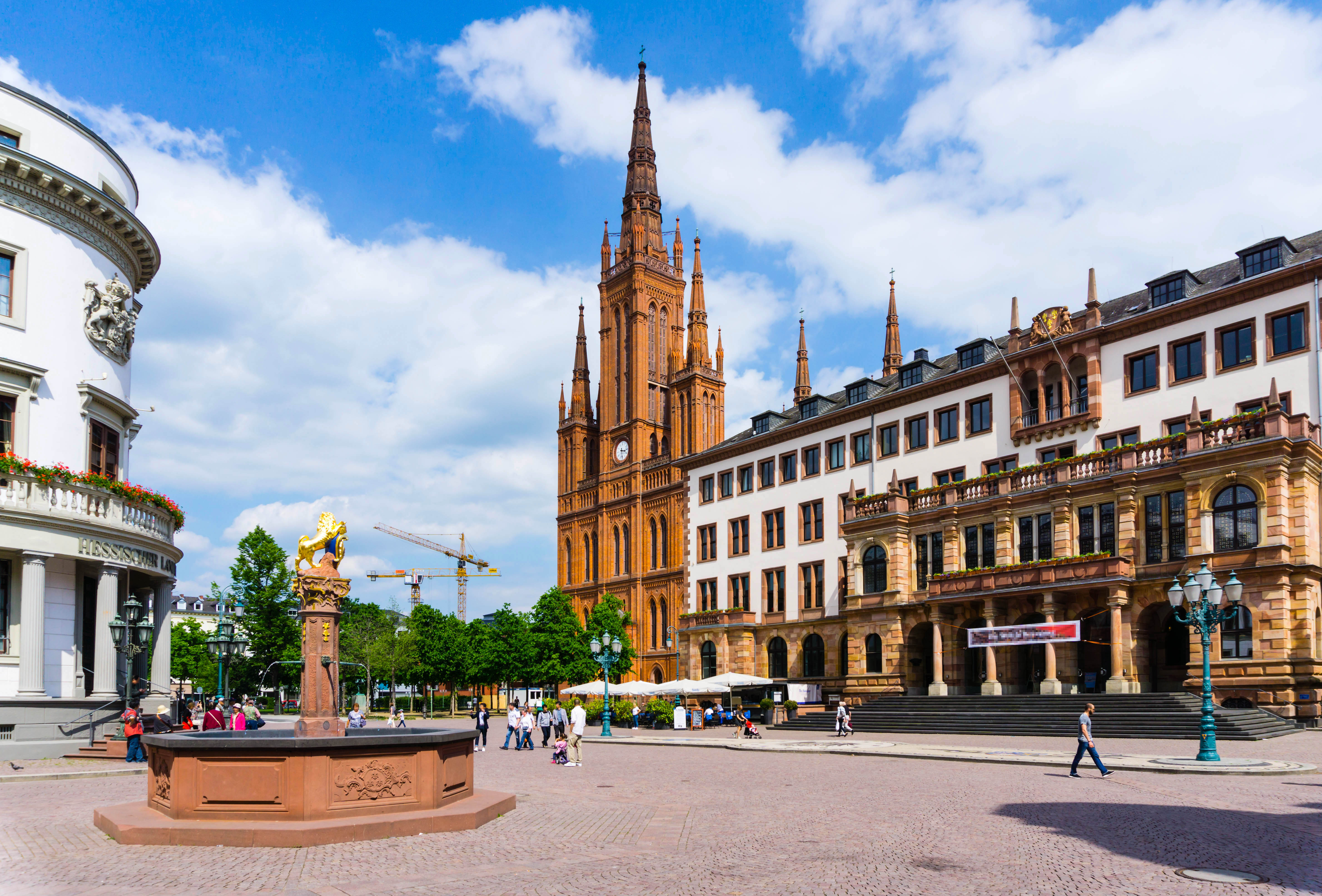 Großer Platz mit Brunnen, historischer Kirche und umliegenden Gebäuden bei Tageslicht.