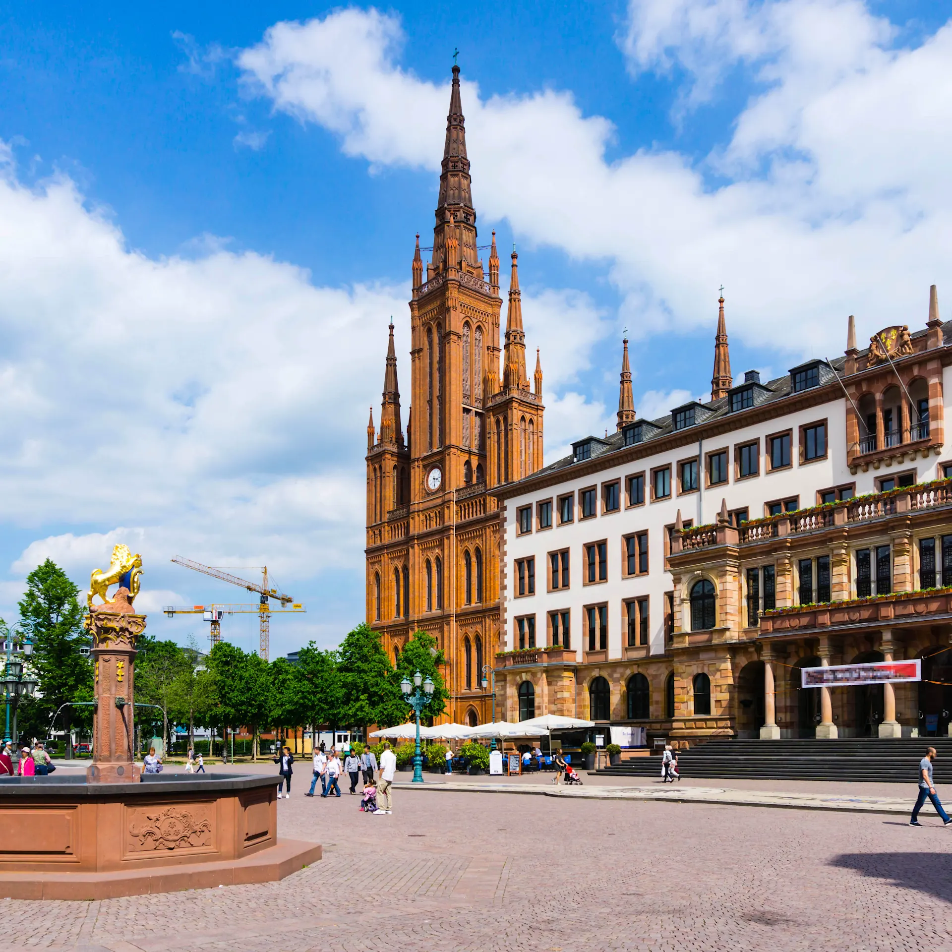 Großer Platz mit Brunnen, historischer Kirche und umliegenden Gebäuden bei Tageslicht.