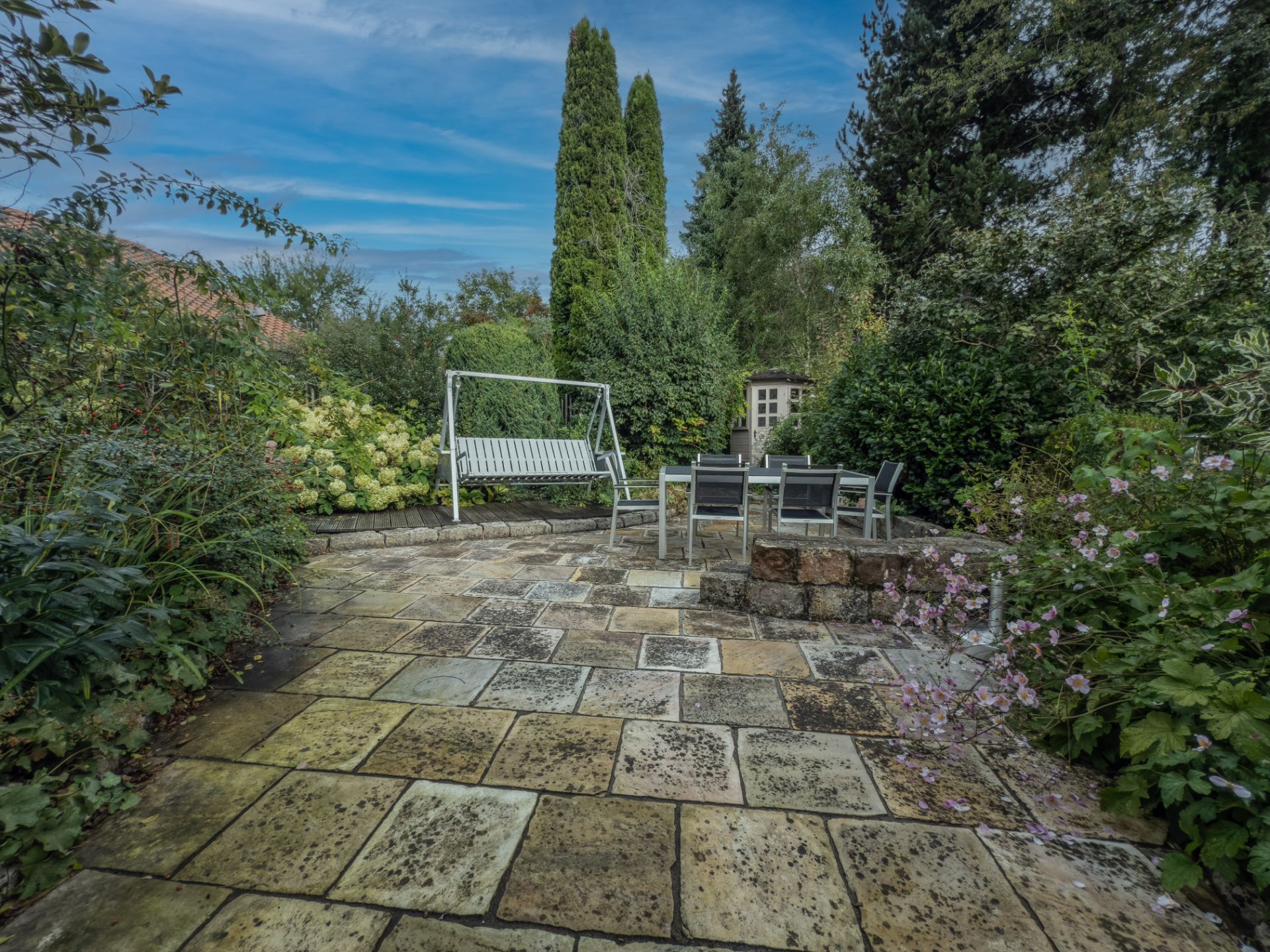 Gepflasterte Terrasse mit Gartenschaukel, Tisch, Stühlen und umgeben von vielen grünen Pflanzen.
