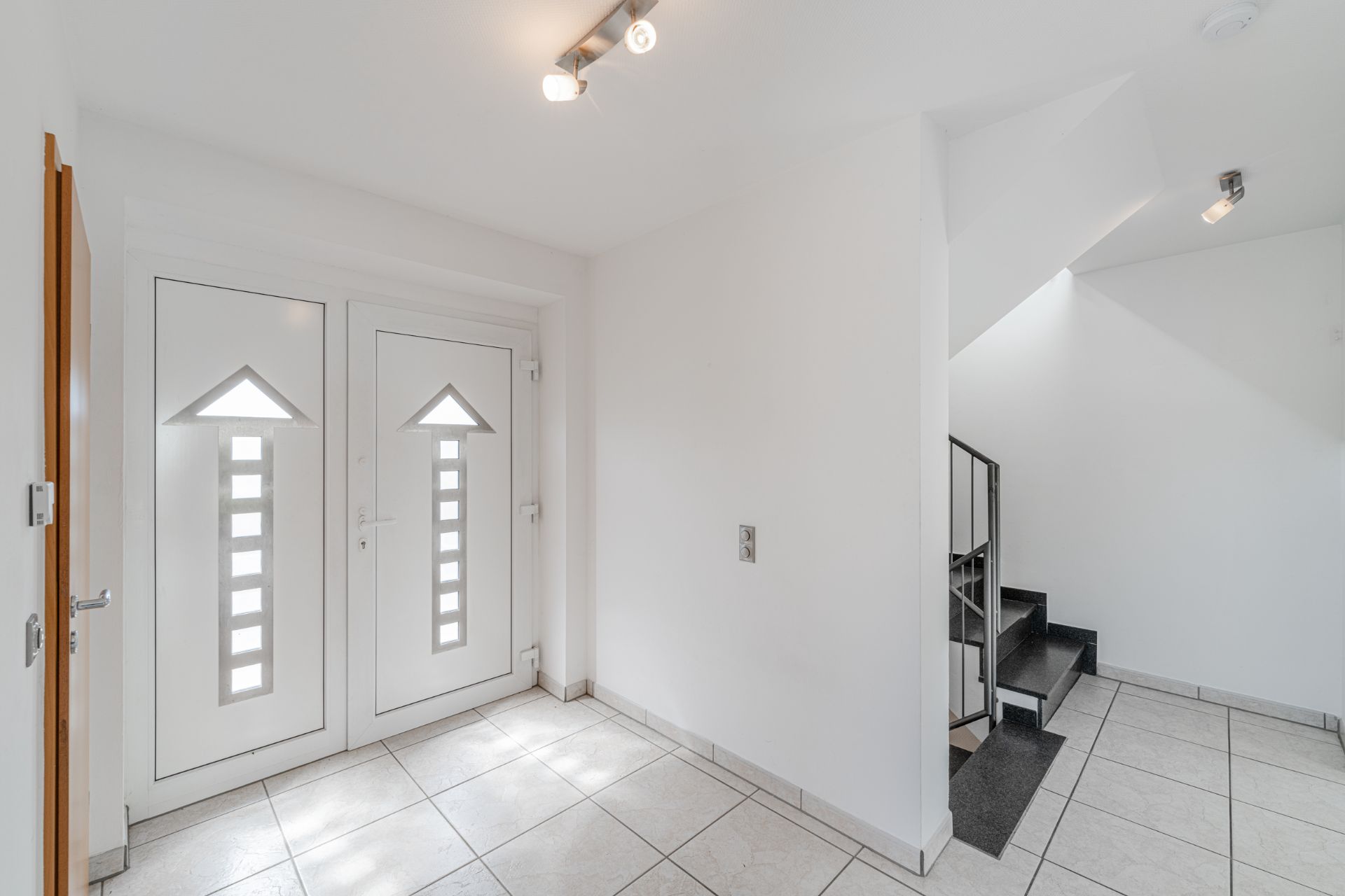 Heller Eingangsbereich mit weißen Türen, Fliesenboden und Treppe mit schwarzem Geländer.